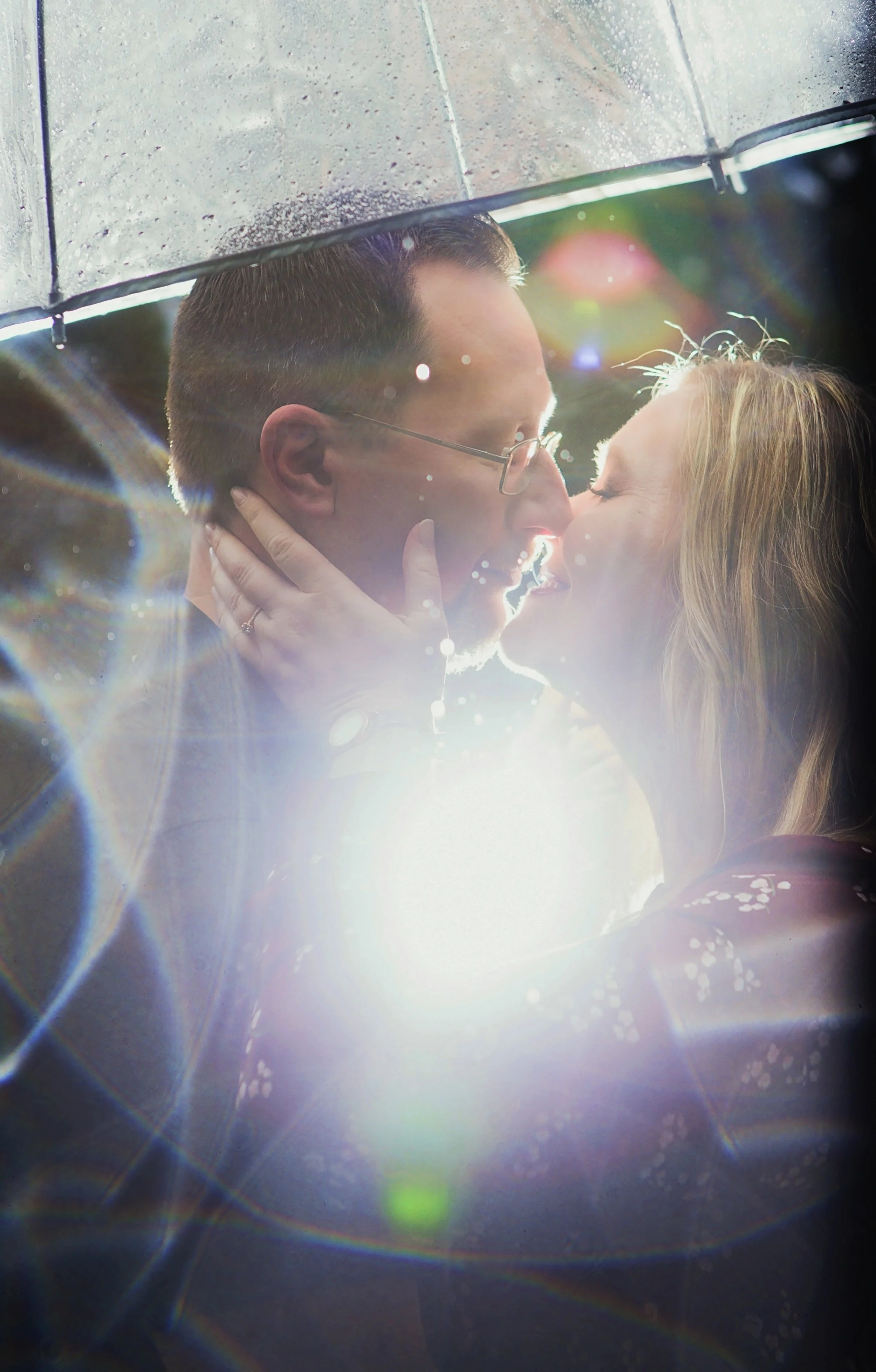 A couple is sharing a romantic kiss under a translucent umbrella on a rainy day, with sunlight creating rainbow lens flares.