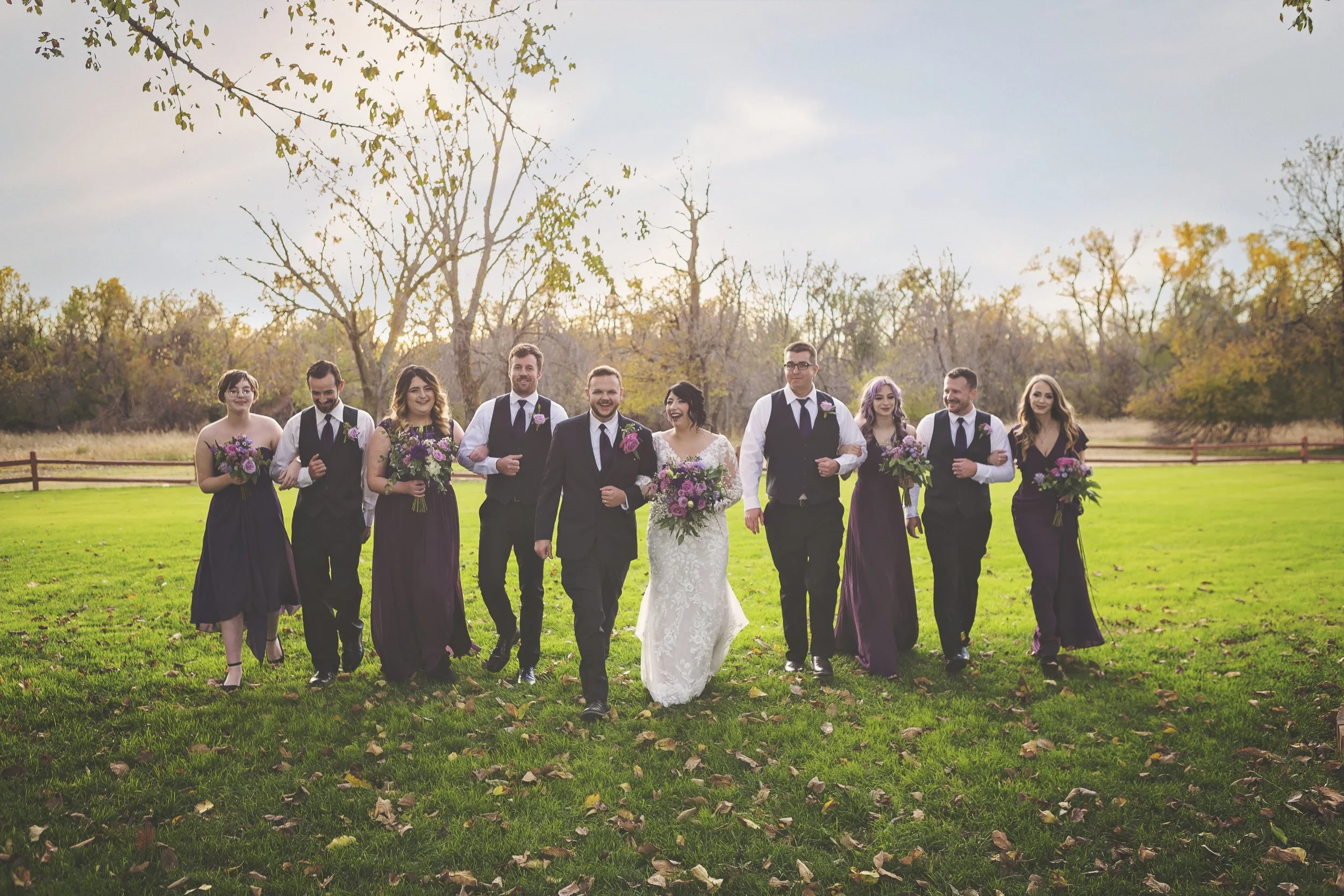 A bride and ten wedding guests walking together on a grassy field during sunset. The bride is wearing a white lace wedding gown and carrying a bouquet, flanked by guests dressed in formal attire, with four women in purple dresses and six men in black