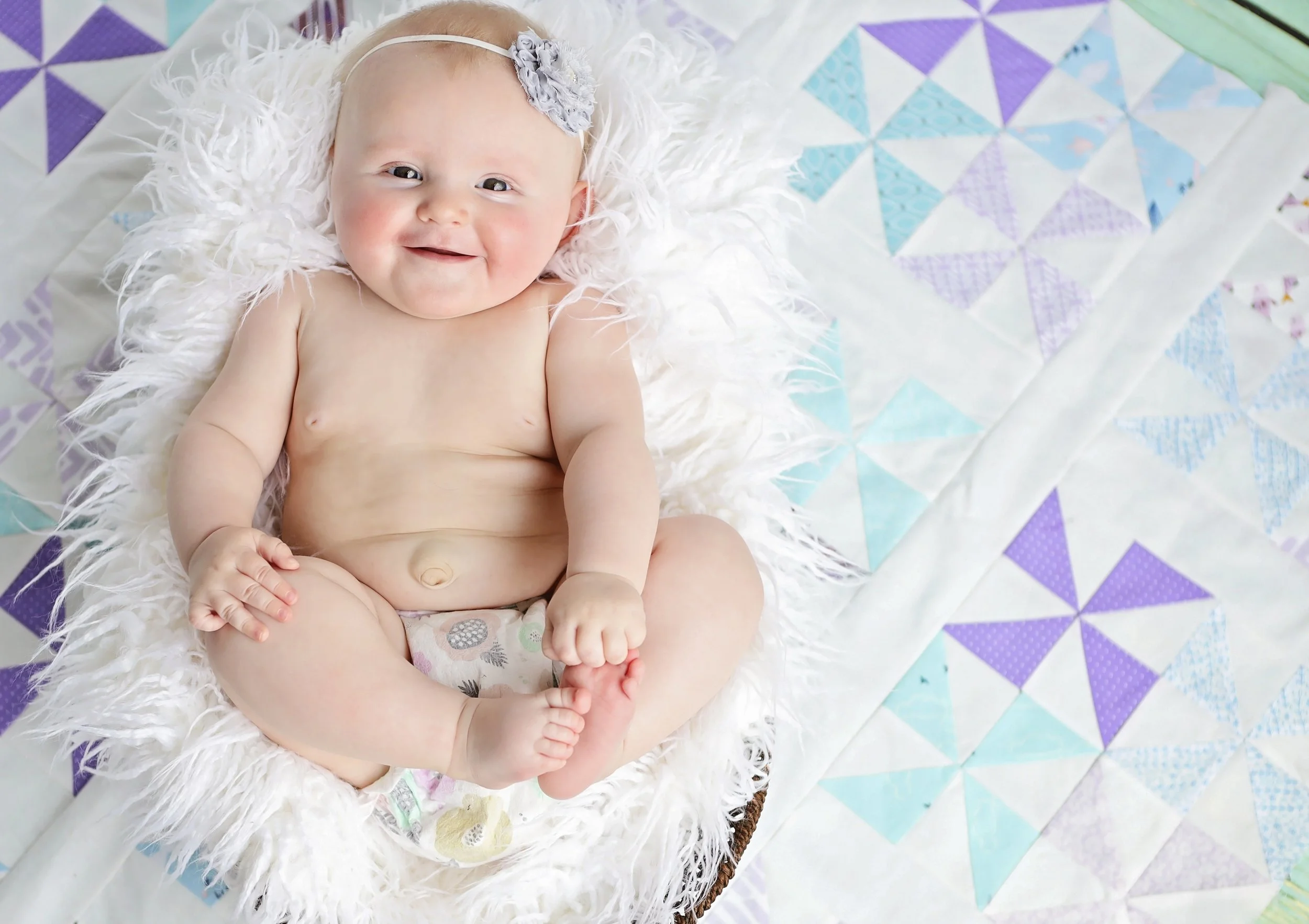 A smiling baby with fair skin, light hair, and a flower headband, sitting on a fluffy white surface on top of a colorful quilt with geometric patterns in purple, blue, and green.