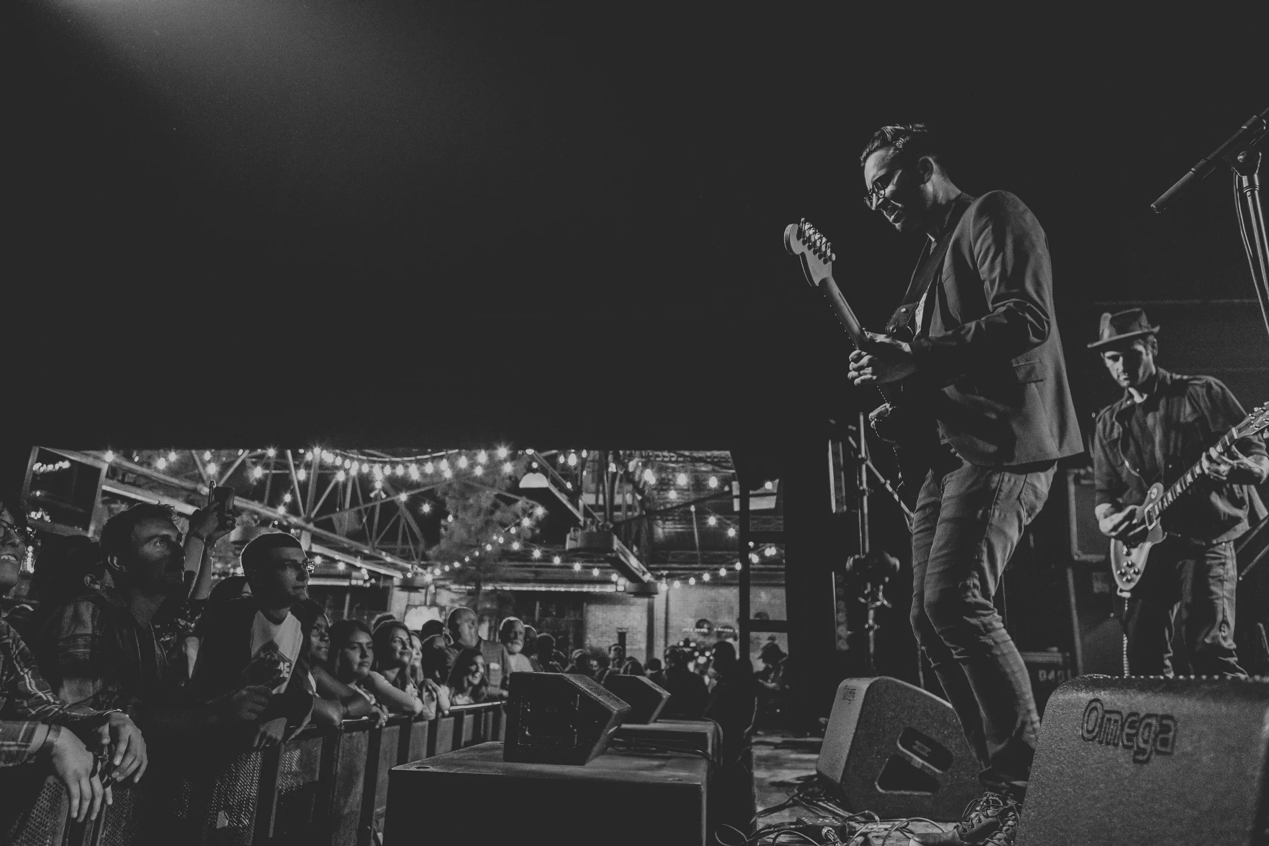Black and white photo of a live music performance at night with a band playing on stage and an audience watching in front of the stage, under string lights.