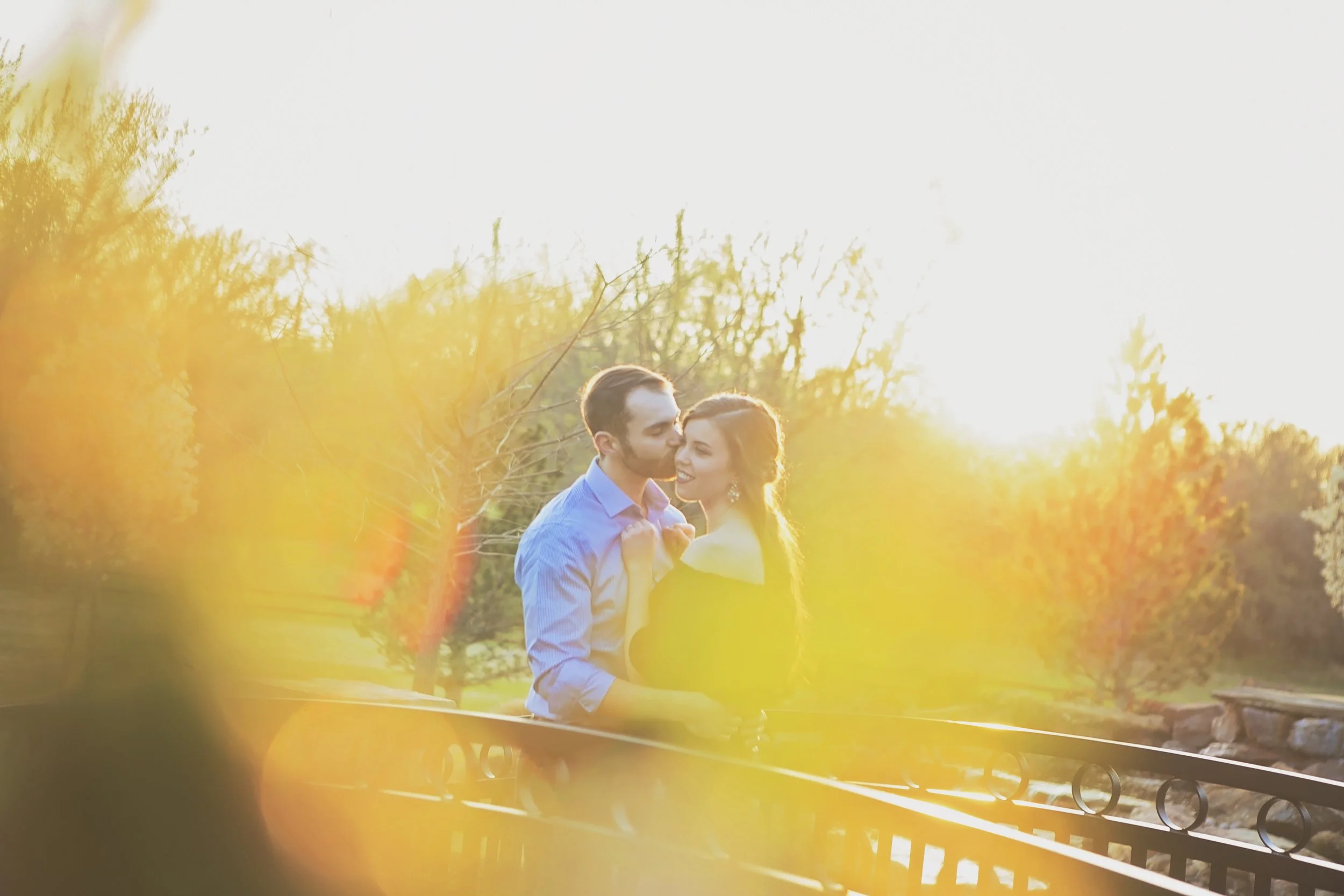 A couple sharing a romantic moment outdoors at sunset, surrounded by trees and foliage.