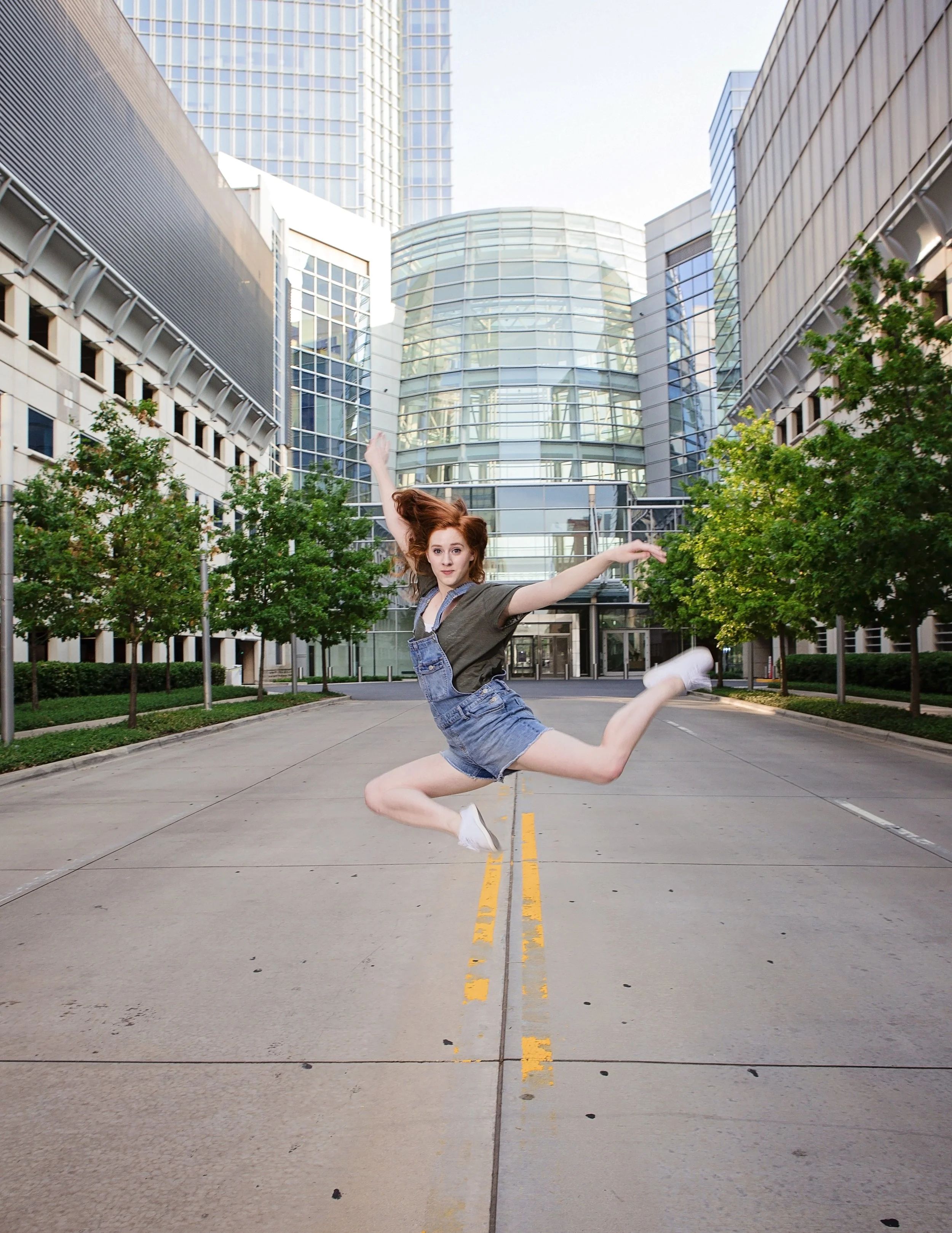 Young woman jumping in the air on an empty city street surrounded by modern glass buildings and trees.