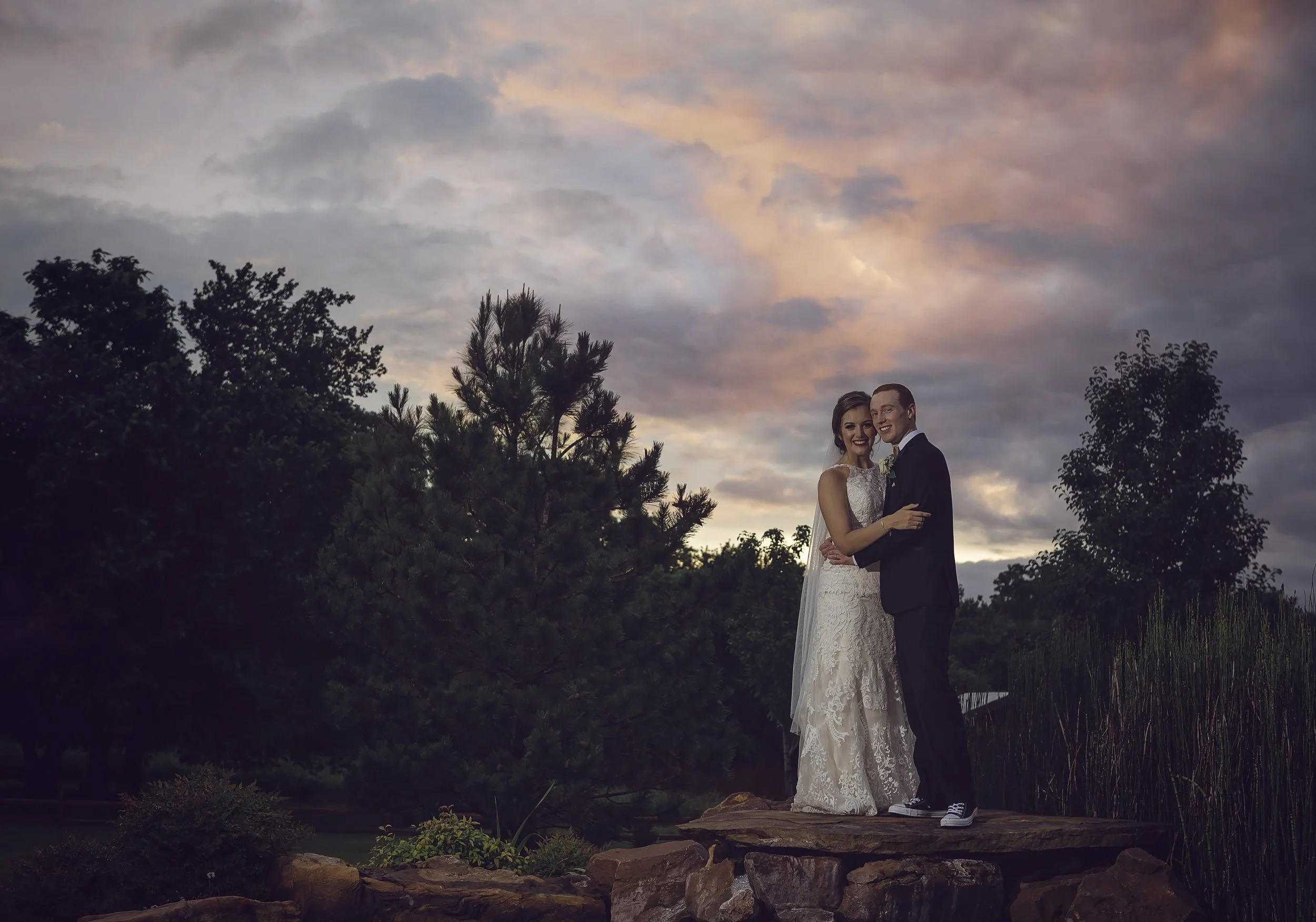 A newlywed couple in wedding attire standing on a large rock outdoors at sunset, with trees and cloudy sky in the background.