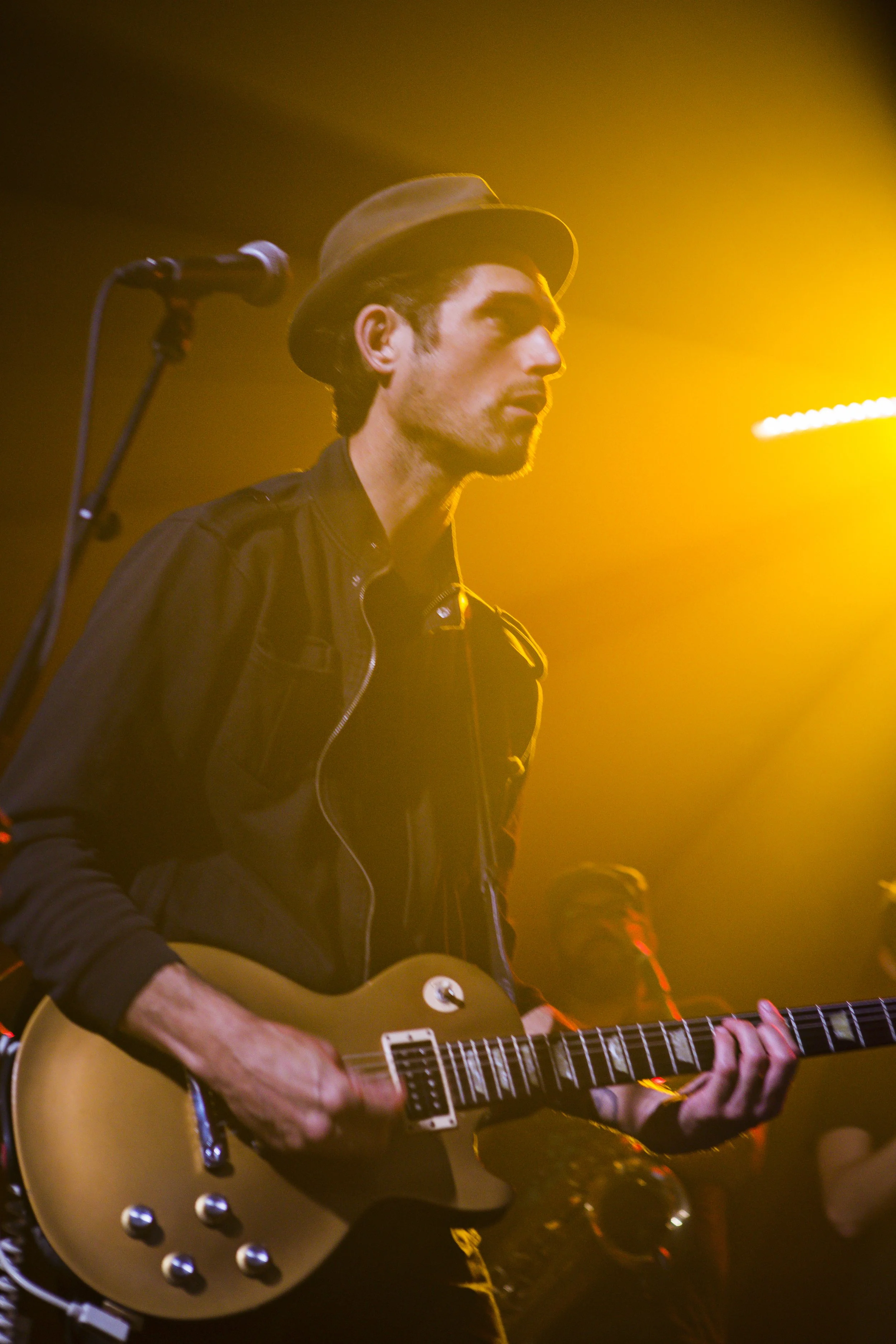A man wearing a brown hat and black jacket playing an electric guitar on stage with warm yellow lighting.