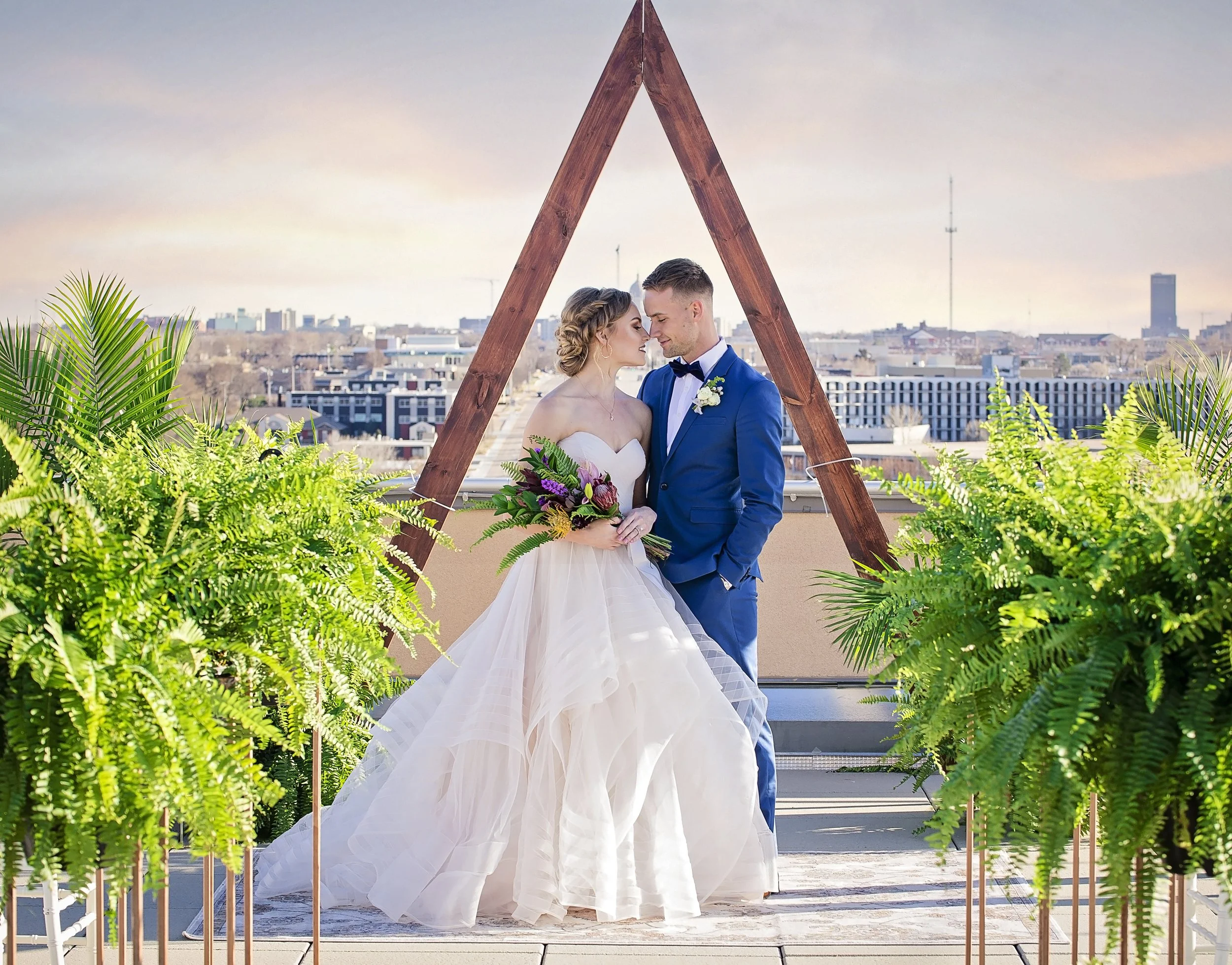 A bride and groom standing close together, nose-to-nose, under a wooden triangular wedding arch on a rooftop, with city buildings in the background, surrounded by green plants.