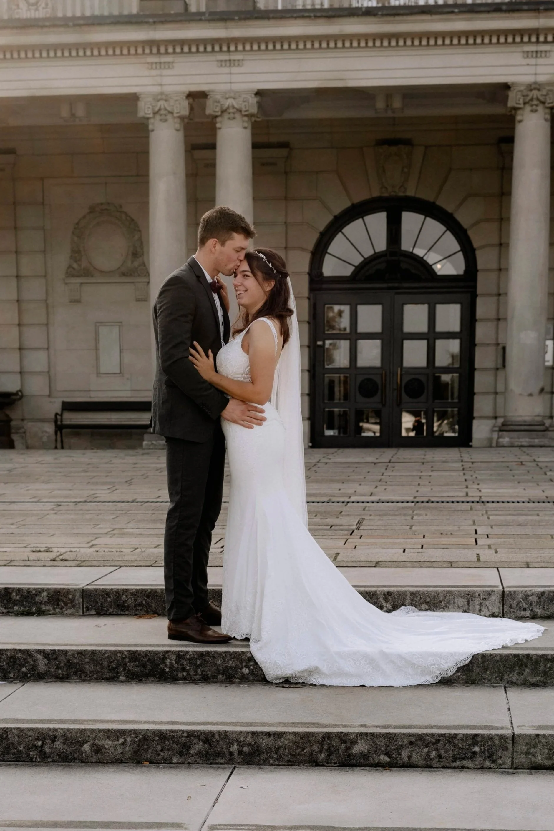 Ein Brautpaar steht vor einem historischen Gebäude, lächelt und hält sich an den Händen. Der Mann trägt einen schwarzen Anzug, die Frau ein weißes Hochzeitskleid. Es ist eine Hochzeitsaufnahme im Freien.