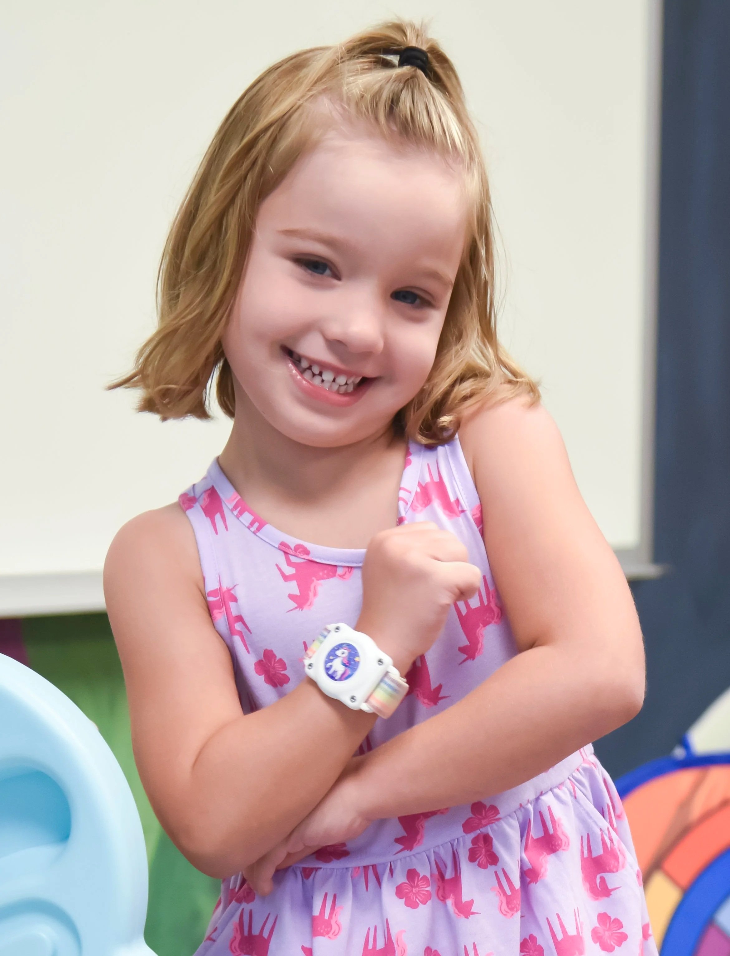 A young girl with light brown hair wearing a pink and purple dress and a rainbow-themed watch, smiling and showing her teeth, standing in front of colorful playground equipment.