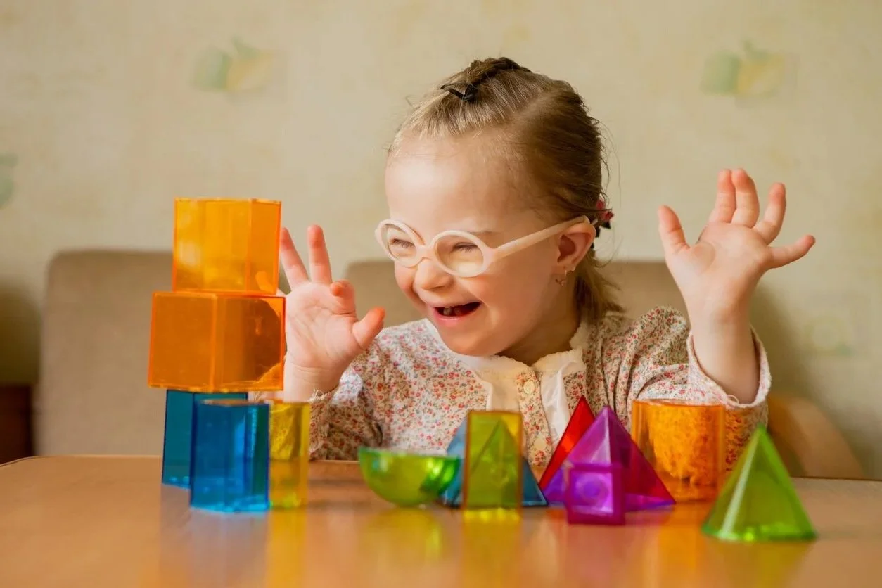 A young girl with glasses and a floral shirt enjoying playing with colorful translucent geometric blocks at a table.