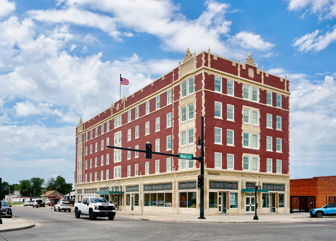 A large red brick historic building with cream accents on the corner of Main Street and another street, with an American flag on top, cars parked in front, and a cloudy sky overhead.