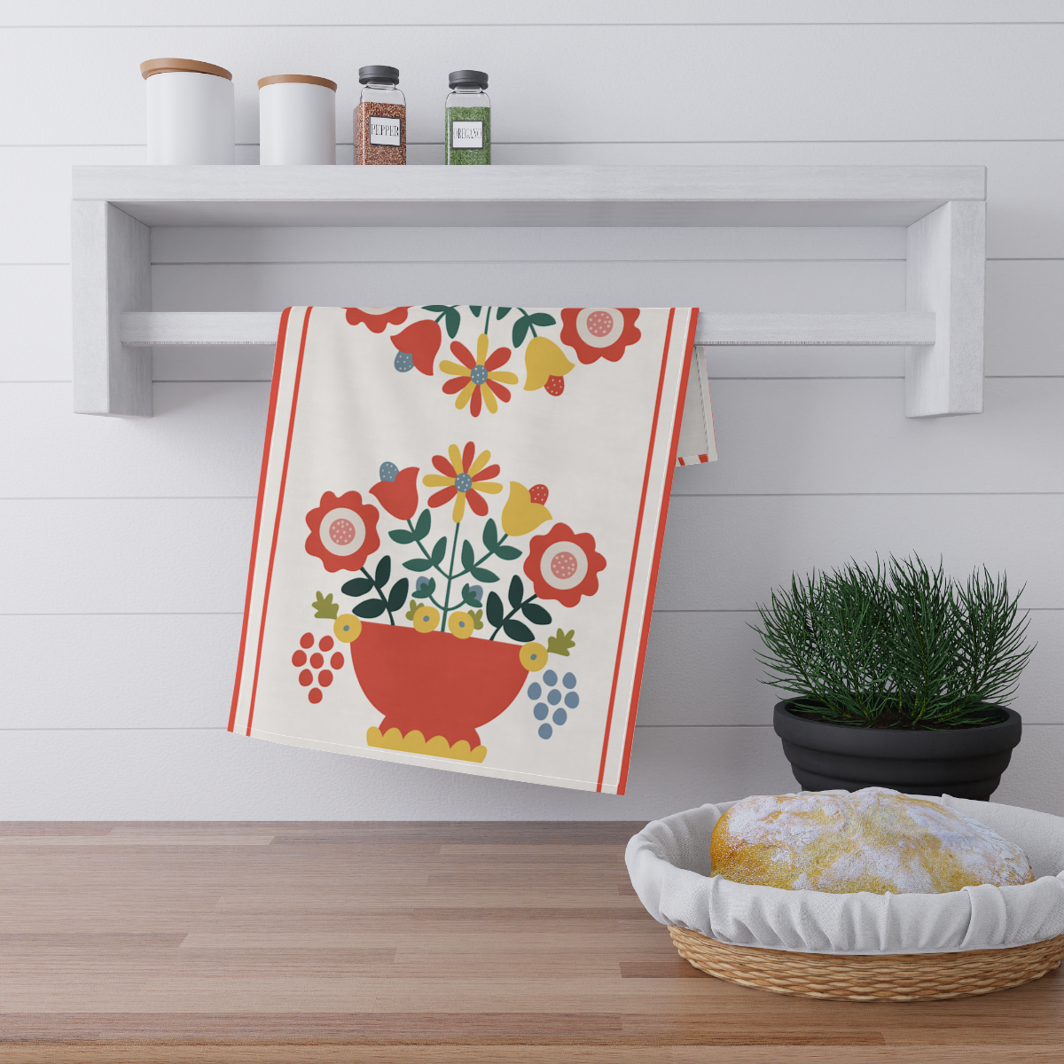 Kitchen scene with shelf and decor. Shelf holds two white canisters and two spice jars labeled "Pepper" and "Herbs." A decorative towel with a colorful floral design hangs below. A black pot with green plant sits below, next to a basket with a round loaf of bread.