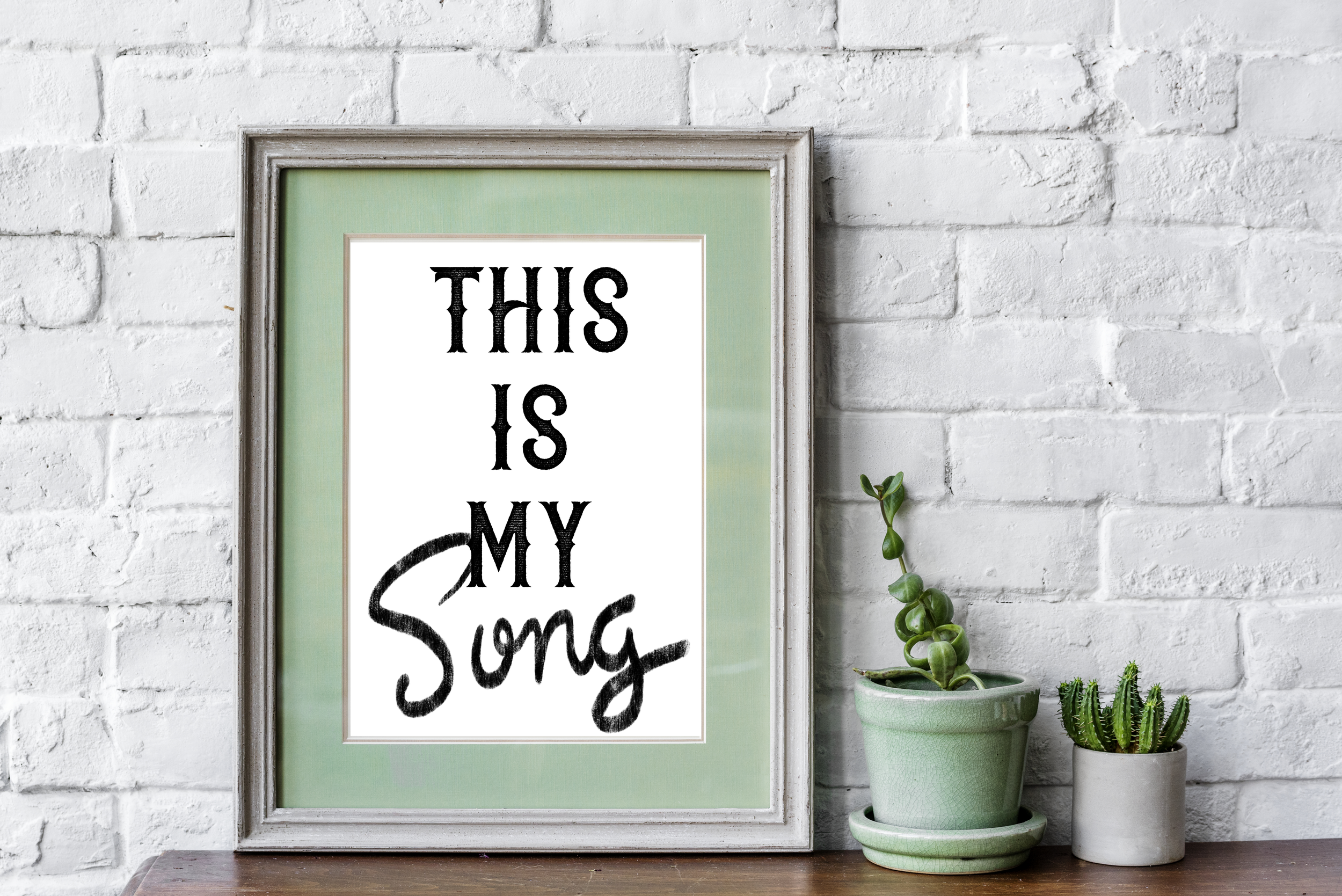 Framed quote "This Is My Song" next to two potted plants on a wooden surface against a white brick wall.