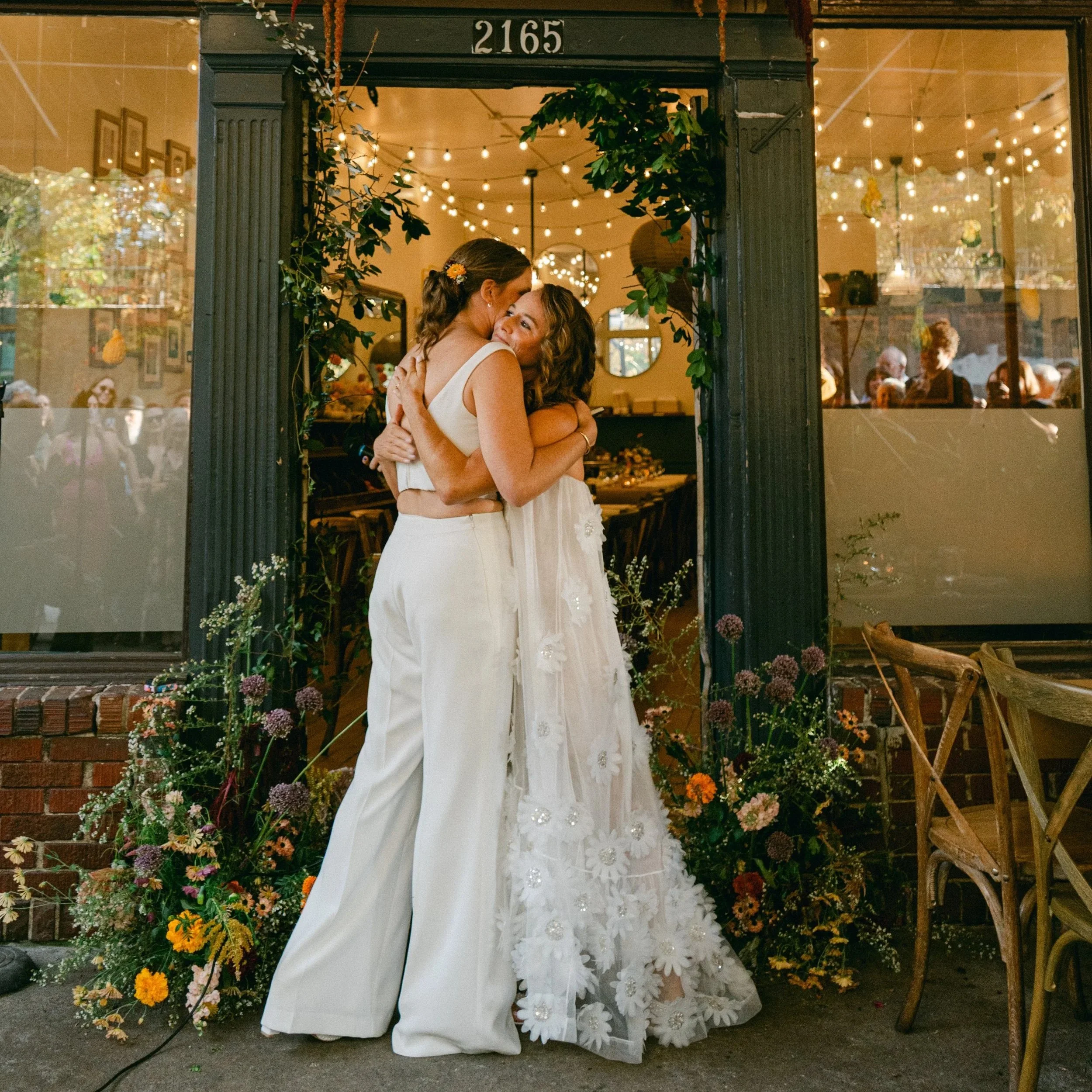  Two brides in white embracing after saying I do. Garden florals frame the altar. A fall wedding at Sun In My Belly in Atlanta, Ga.