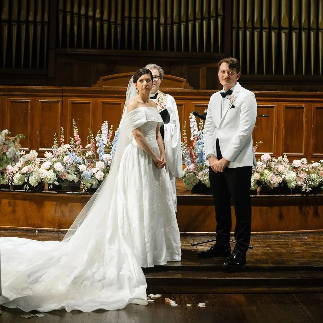 A wedding ceremony taking place inside a church with a stained glass window depicting religious figures. The bride in a white gown with a veil and the groom in a white tuxedo stand before a pastor, surrounded by floral arrangements.