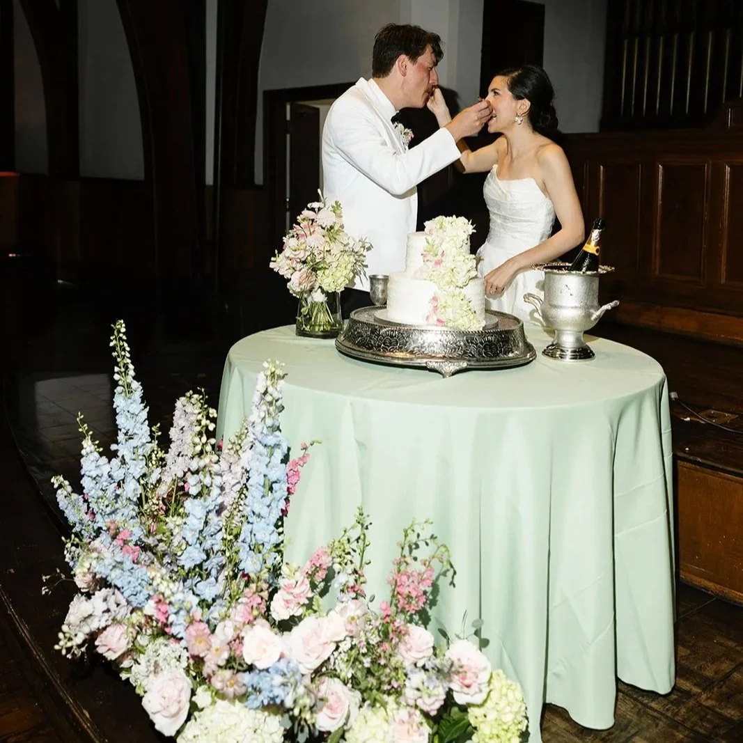 A bride and groom are celebrating their wedding with a cake on a decorated table. The groom is feeding the bride, who is smiling, in a dimly lit room with wooden ceiling beams and floral arrangements in the foreground.