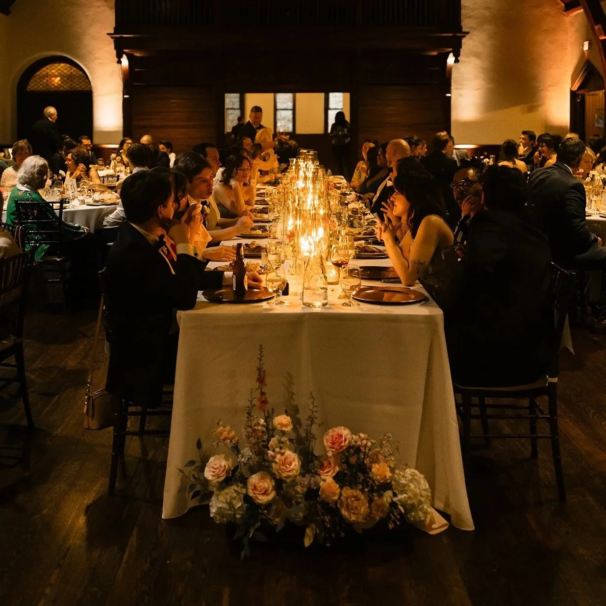 A formal dinner gathering in a large, dimly lit room with a vaulted ceiling and hanging lanterns. Guests are seated at long tables, enjoying a meal with candles and floral centerpieces.
