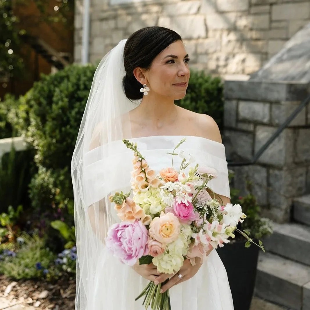 Bride with dark hair in a bun, wearing a white off-shoulder wedding dress, holding a bouquet of pink and white flowers, standing outside near a stone building with garden greenery.