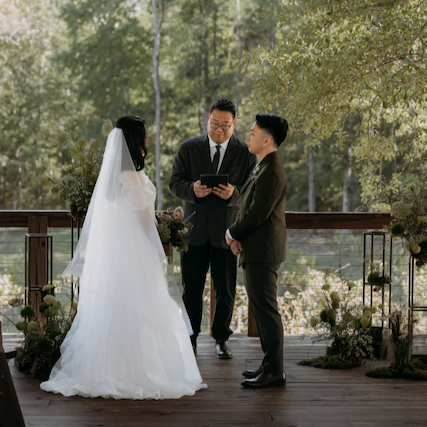 A wedding ceremony with a bride in a white dress and veil, a groom in a suit, and an officiant holding a book, outdoors on a wooden platform decorated with flowers, surrounded by trees.