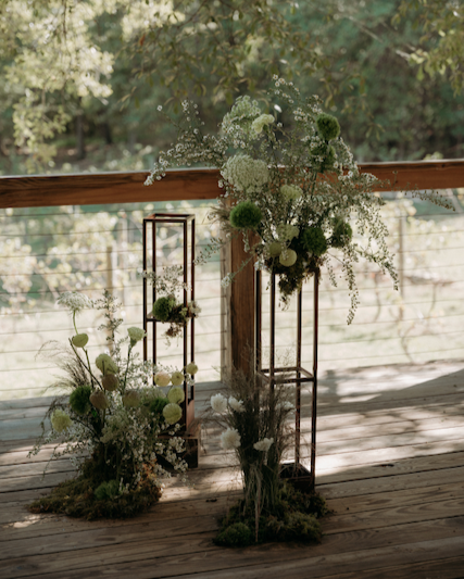Organically styled wedding ceremony arrangements with white and green flowers on wooden stands at an outdoor wedding ceremony and trees in the background.
