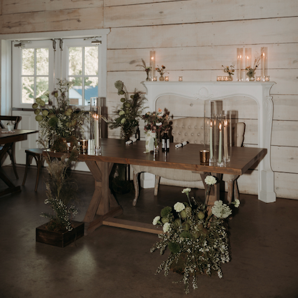 A decorated sweetheart table during the reception, floral arrangements, candles, and a fireplace with candles on top, in a room with large windows and white walls.