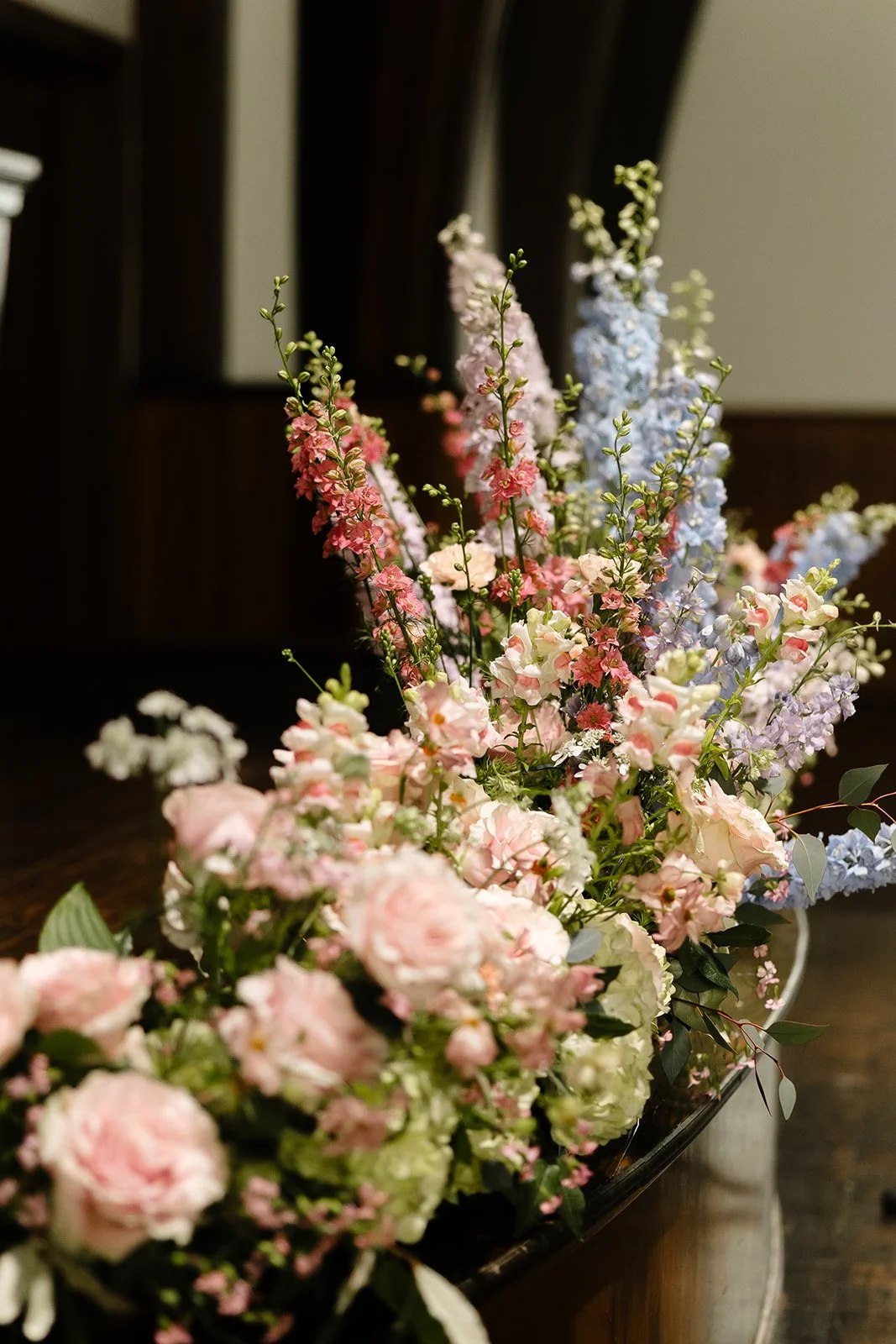 A close-up of a colorful flower arrangement with pink, white, purple, and blue flowers in a vase on a dark wooden table.