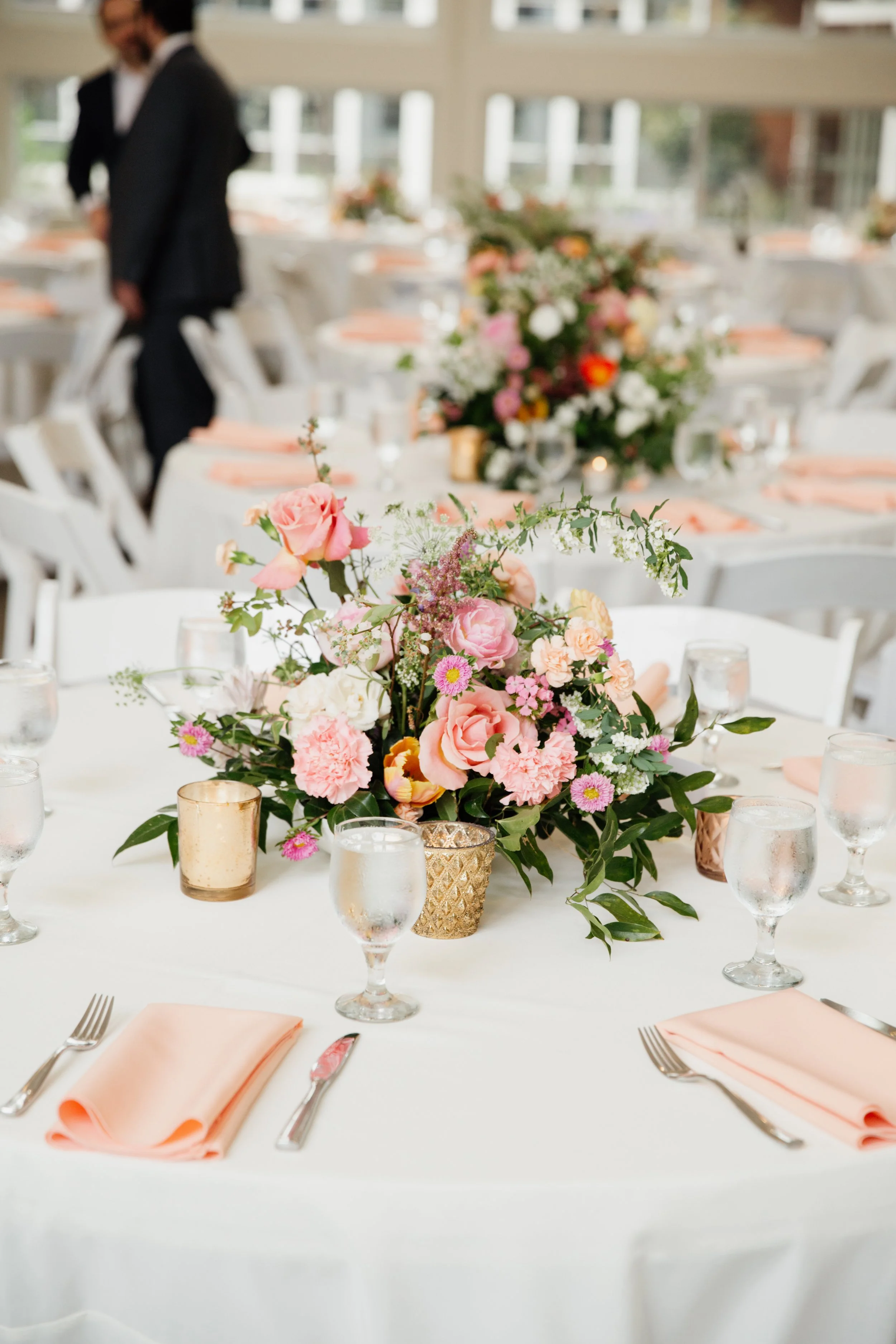 A decorated banquet table with a pink floral centerpiece, surrounded by water glasses, gold votive candles, pink napkins, and silverware, set for a formal event indoors with windows in the background.