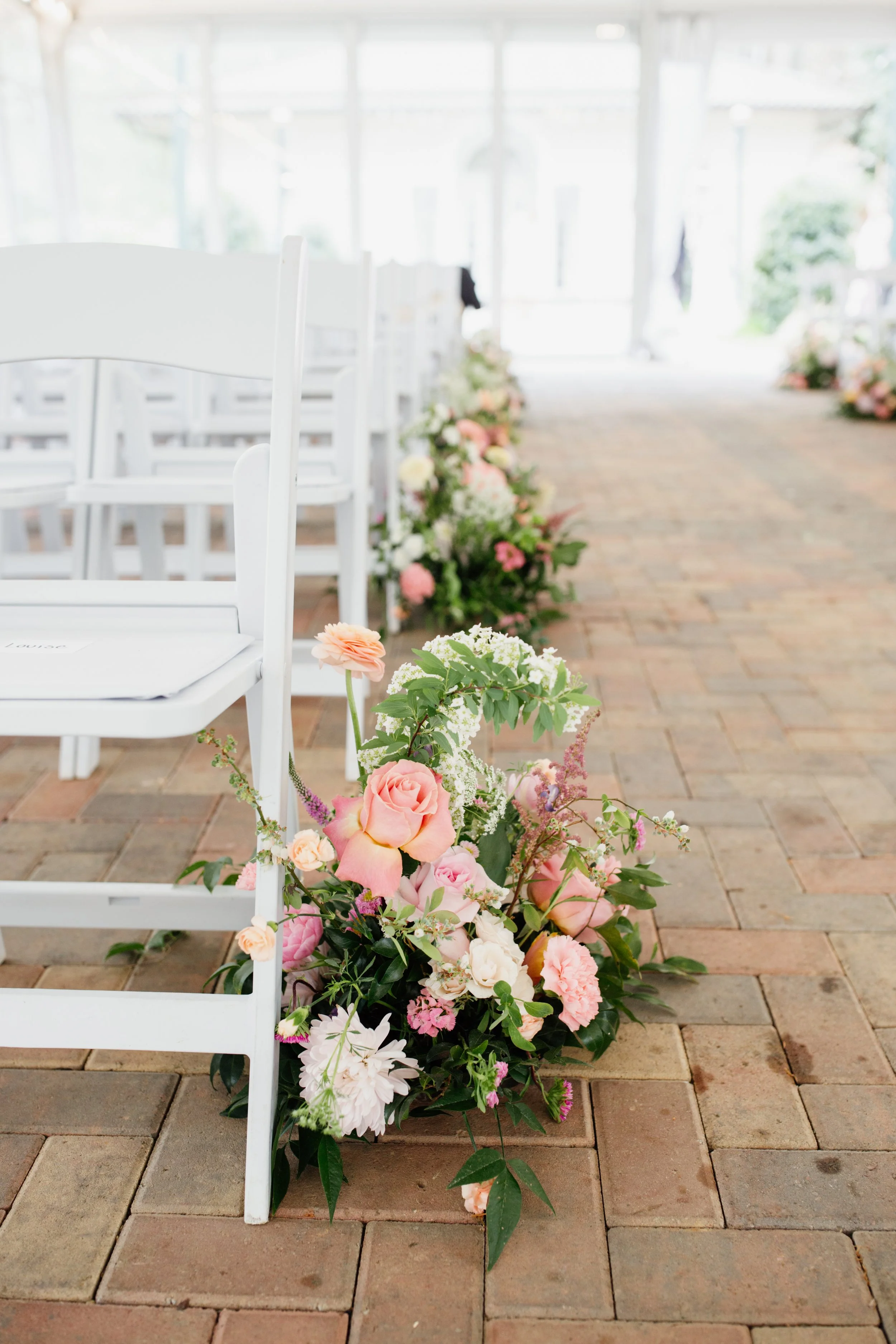 Rows of white chairs with floral arrangements along the aisle of an indoor wedding venue.