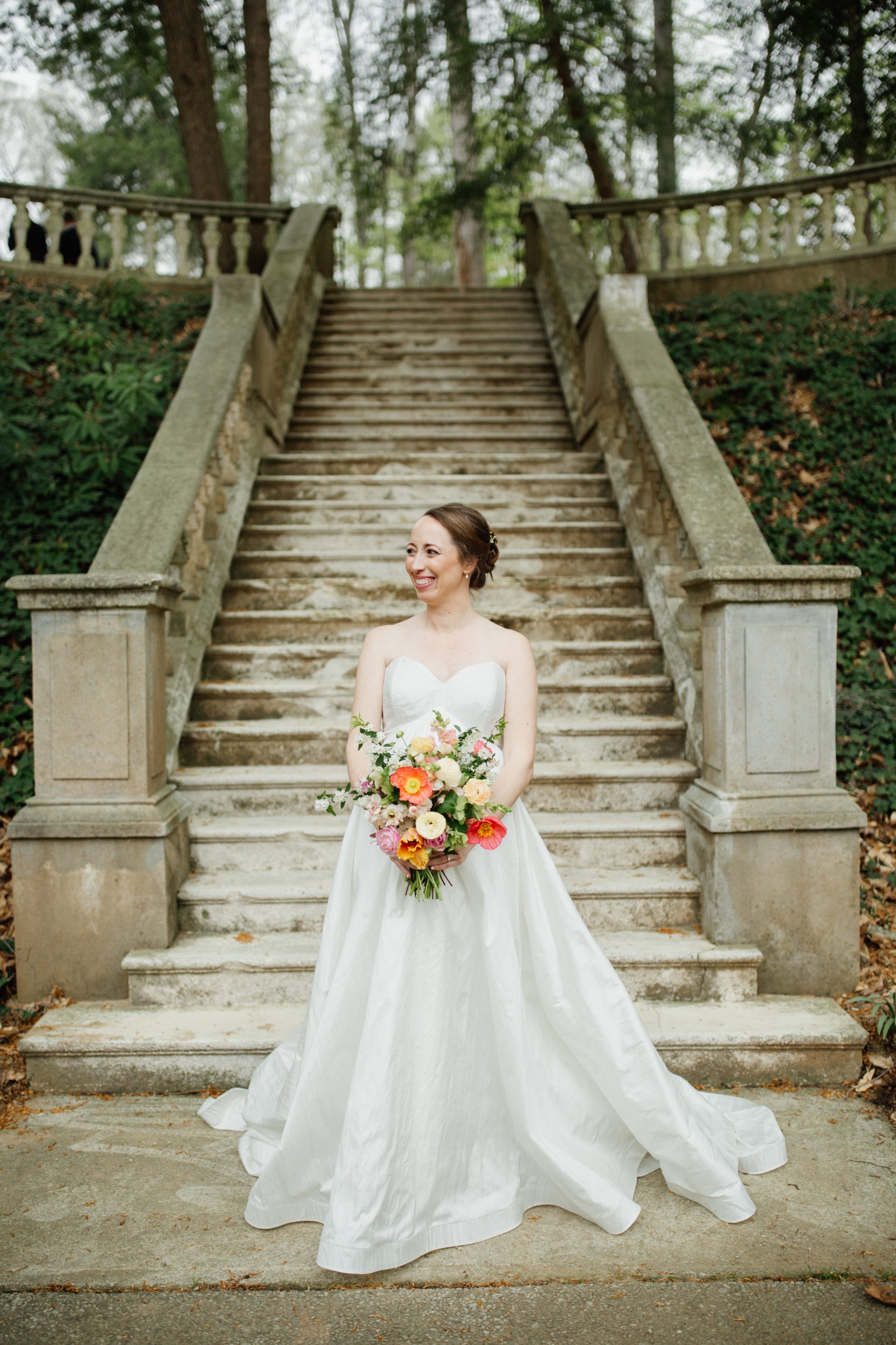 A bride in a white wedding dress holding a colorful bouquet stands at the bottom of outdoor stone stairs surrounded by greenery.