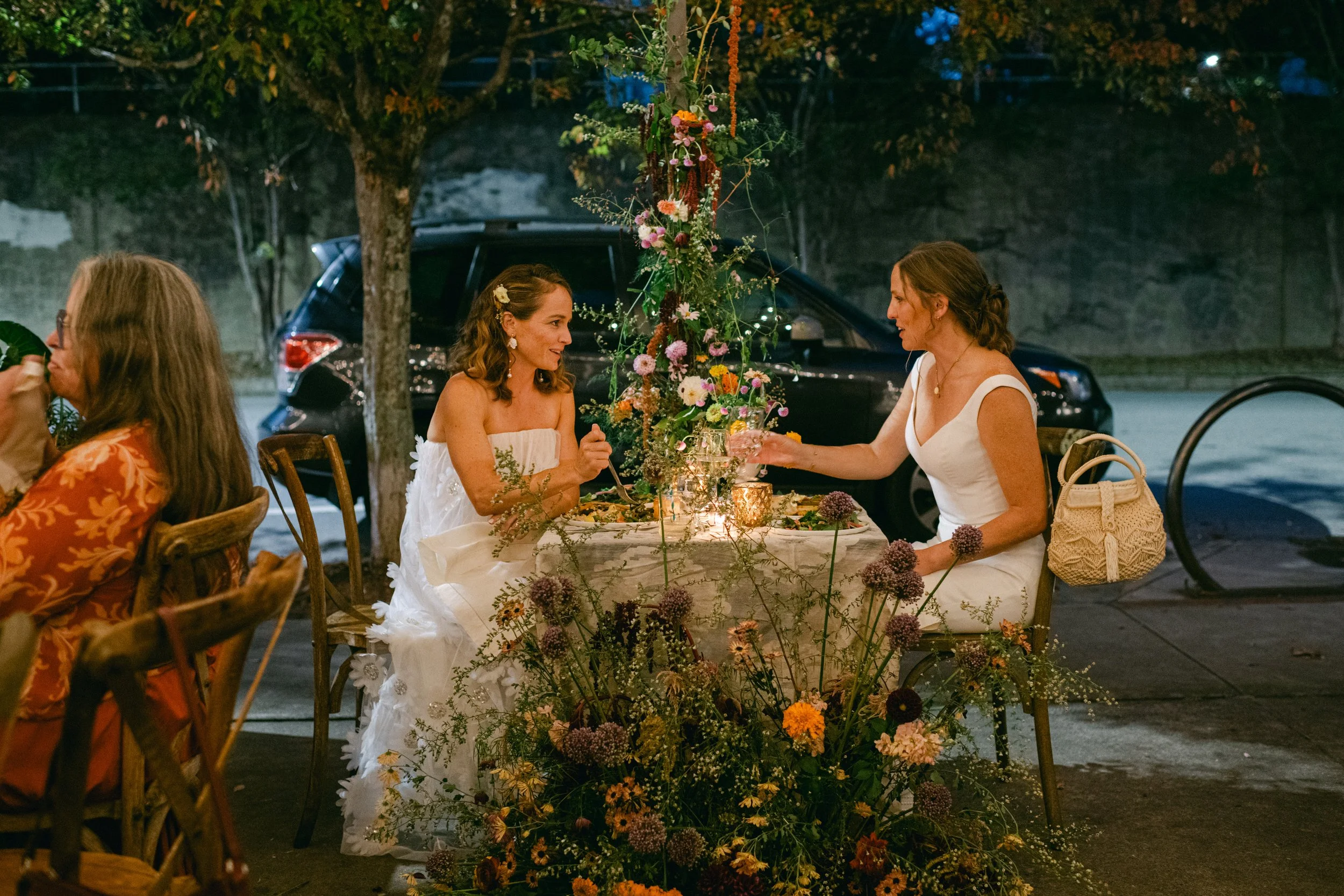 Two brides sit at a decorated sweetheart table with flowers, sharing a moment, during a wedding reception. The setting includes trees, a parked car, and decorative lighting.