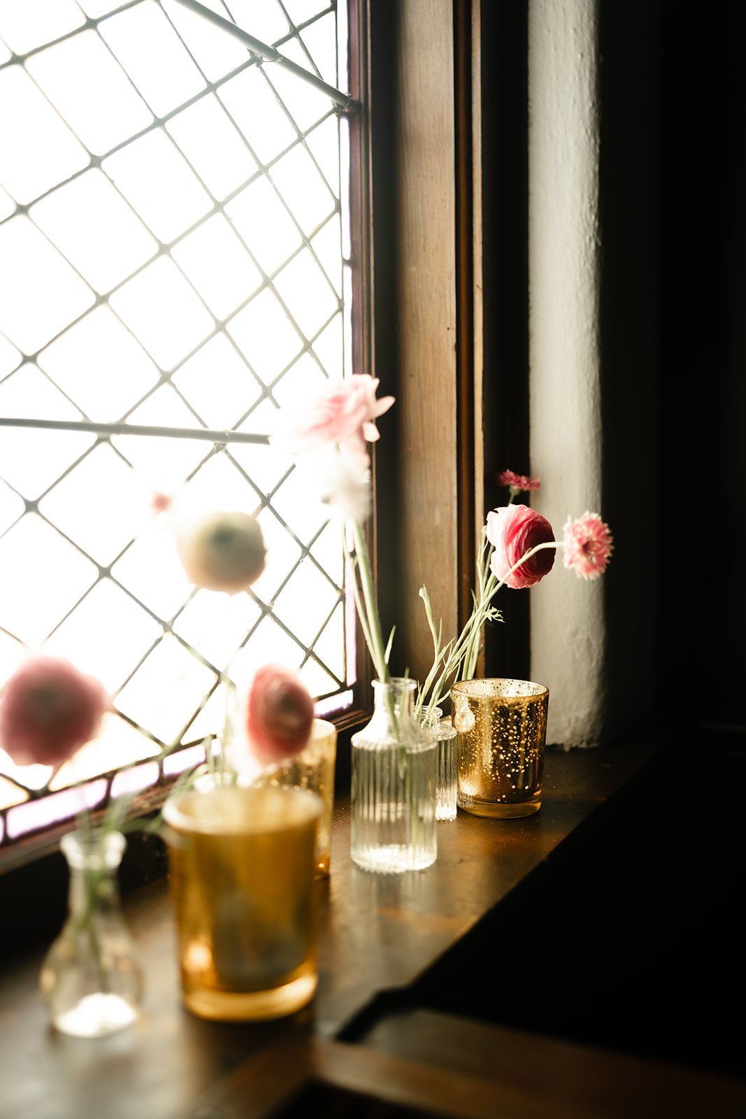 Flowers in glass vases and gold candle holders on a windowsill, with sunlight streaming through a diamond-patterned window.