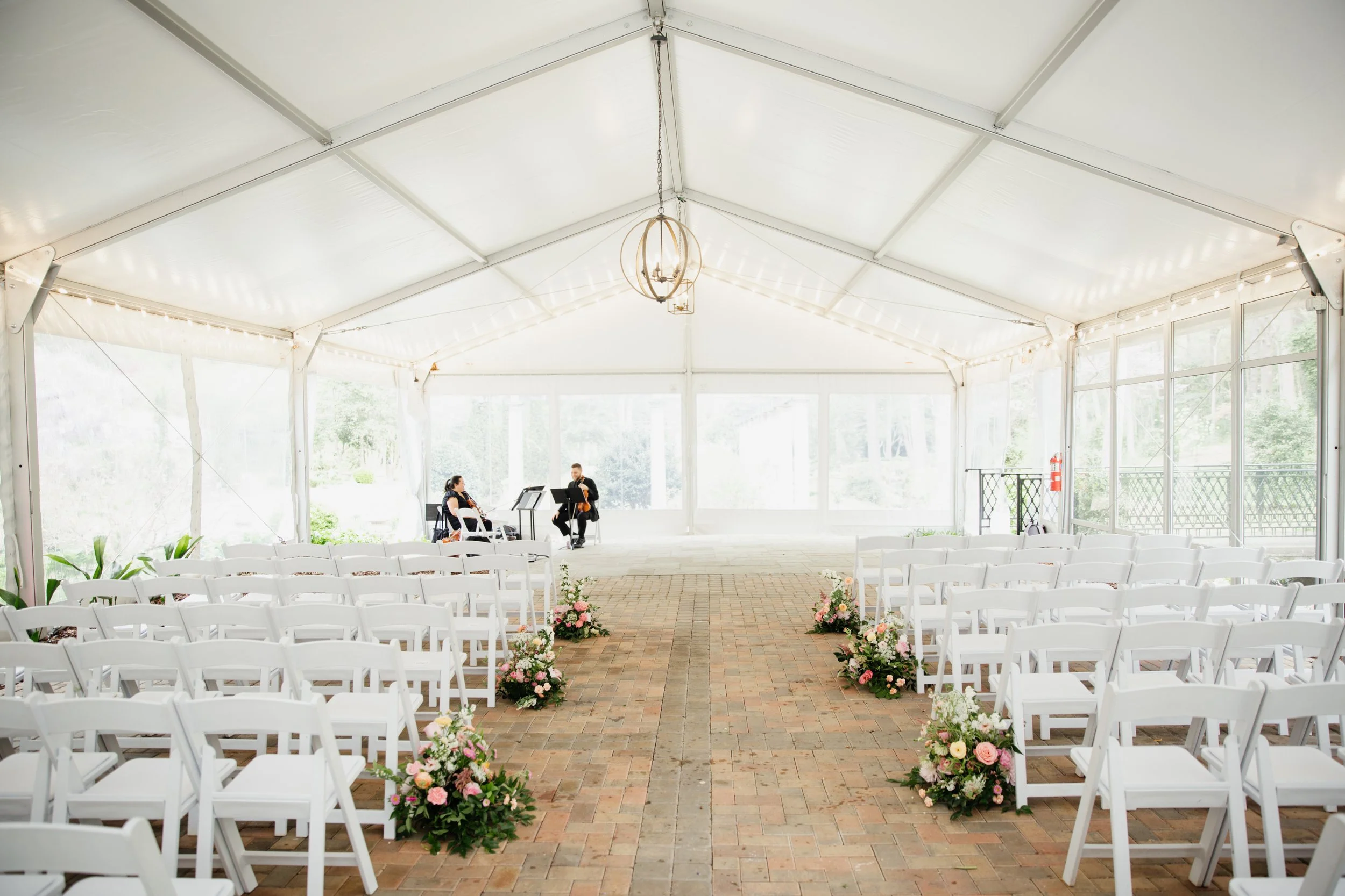 Empty wedding ceremony tent with white chairs, floral arrangements along the aisle, and musicians practicing at the front.