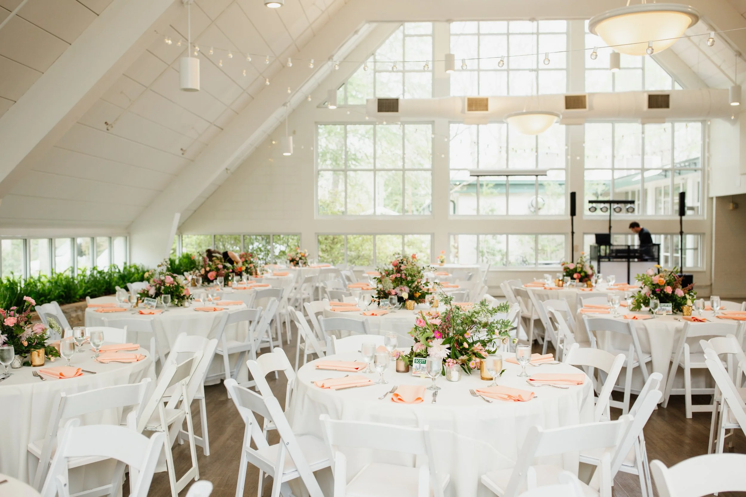 Elegant event space decorated with round tables covered in white tablecloths, pink floral centerpieces, peach napkins, glassware, and silverware, with large windows and indoor lighting.