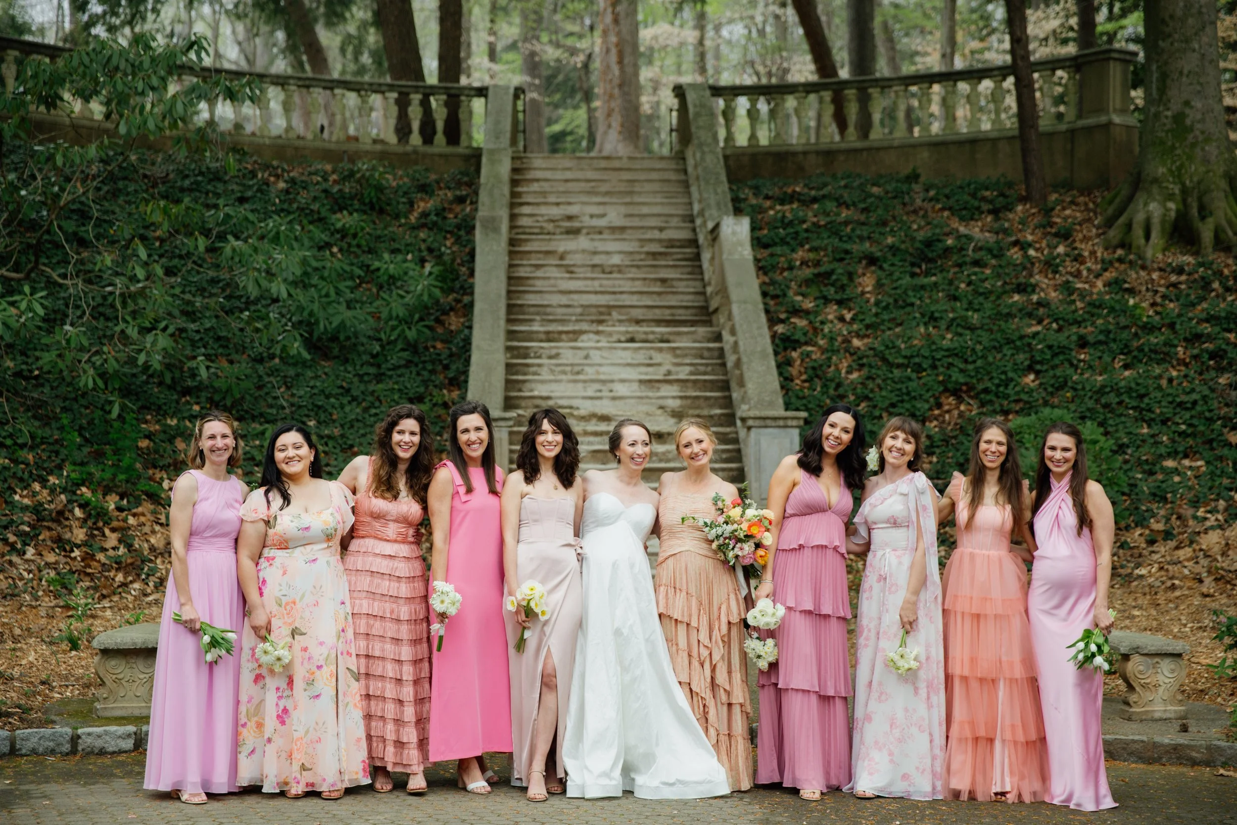 Group of women dressed in pink and pastel-colored dresses, standing outdoors in front of a stone staircase surrounded by greenery.