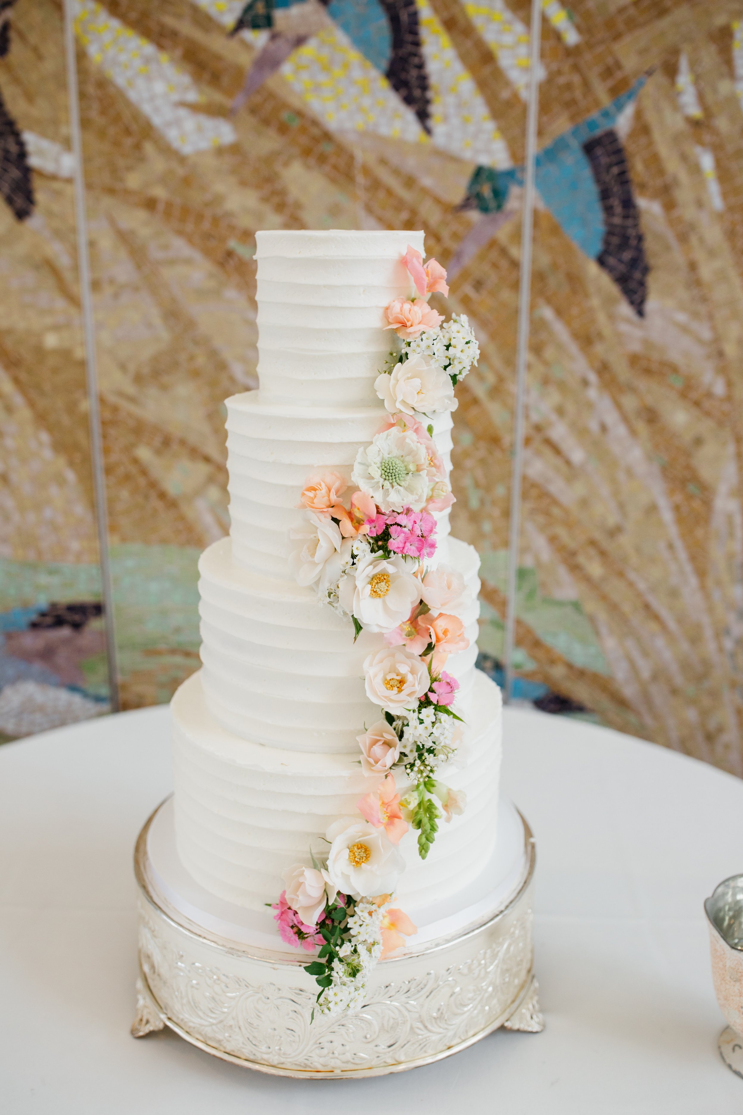 A tall, four-layer white wedding cake with a textured pattern, decorated with cascading pastel-colored flowers, placed on an ornate silver cake stand.