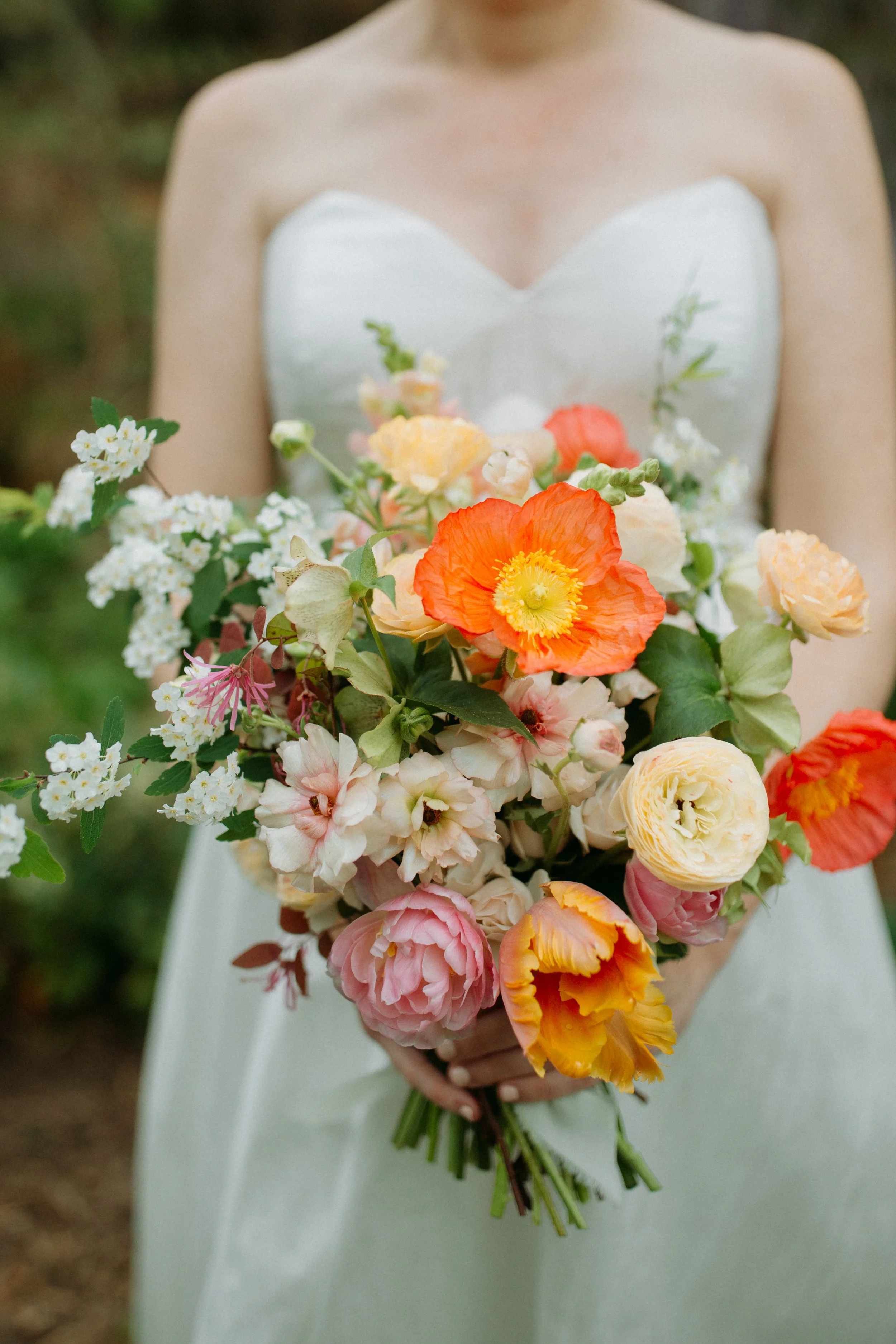 A woman in a strapless white wedding dress holding a large bouquet of colorful flowers, including orange, yellow, pink, and white blooms, outdoors with blurred greenery in the background.