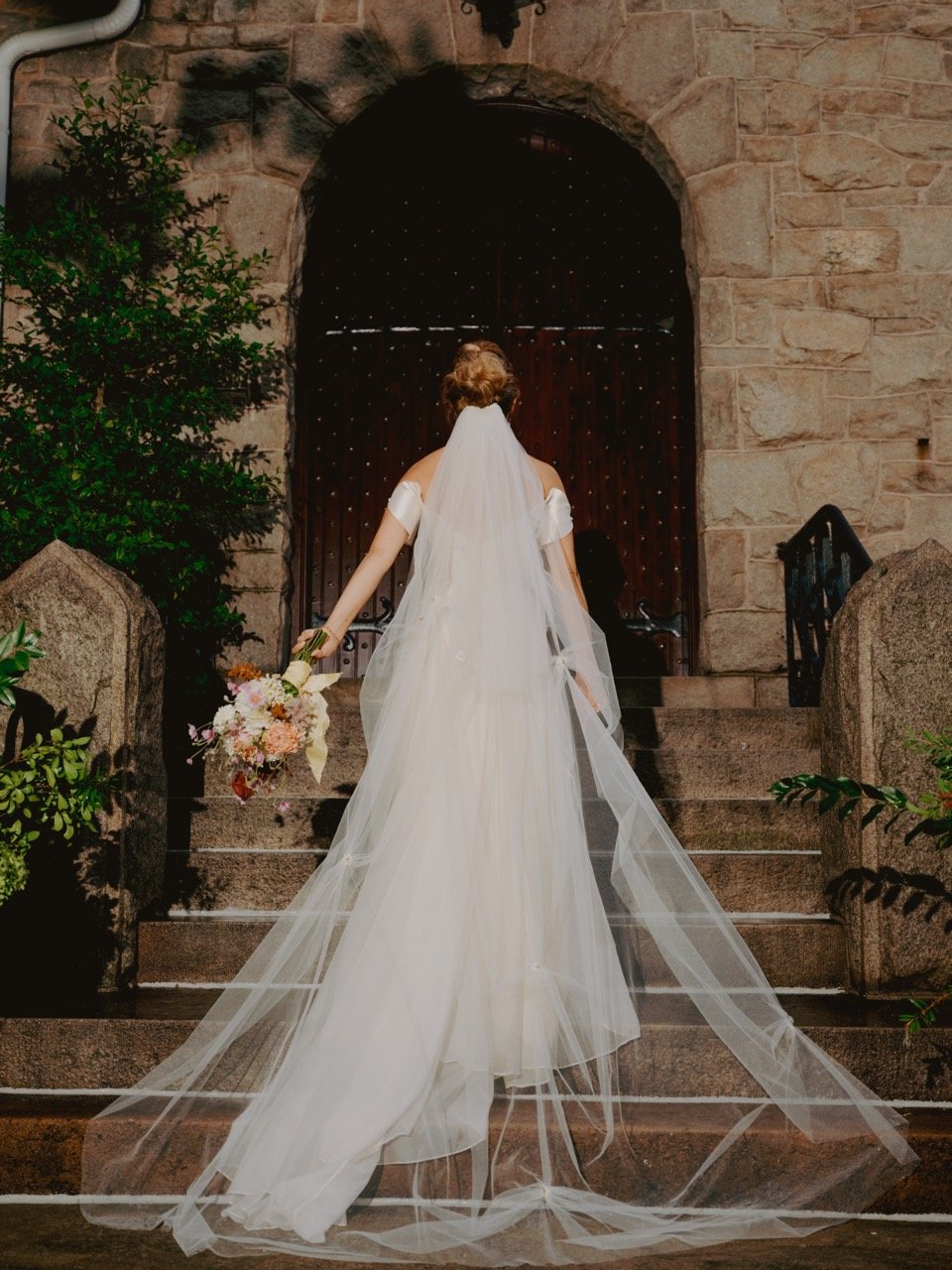 Bride in white dress with veil holding bouquet, walking up stone steps towards arched wooden door of stone building.