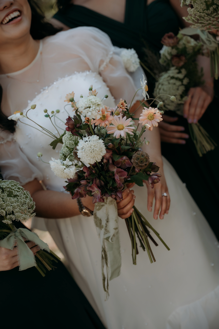 A bride holds a bouquet of fall-inspired flowers, smiling, with a bridesmaid partially visible in the background, also holding flowers.