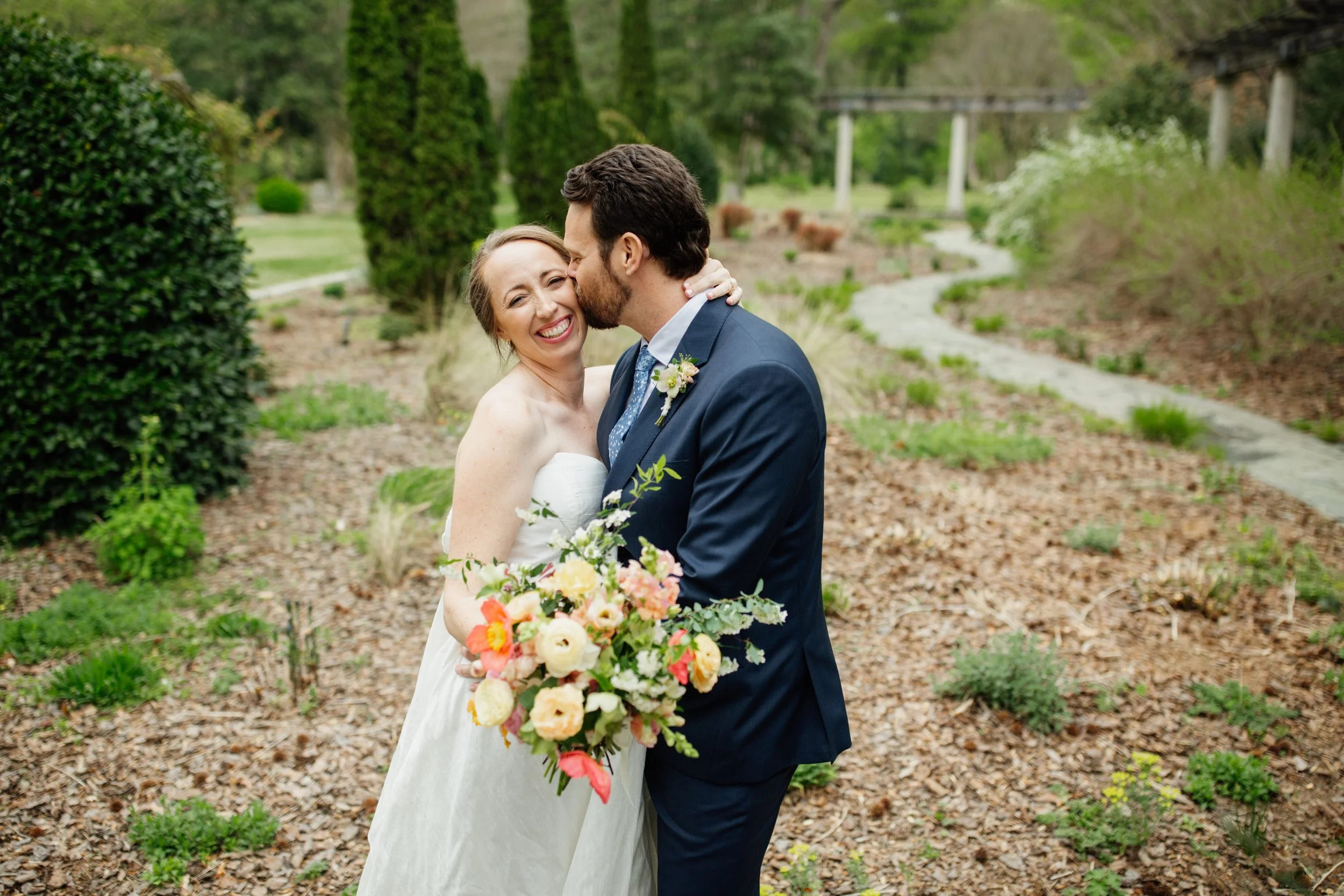 A bride and groom sharing a joyful moment on their wedding day, outdoors in a garden setting, with the bride holding a colorful bouquet of flowers.