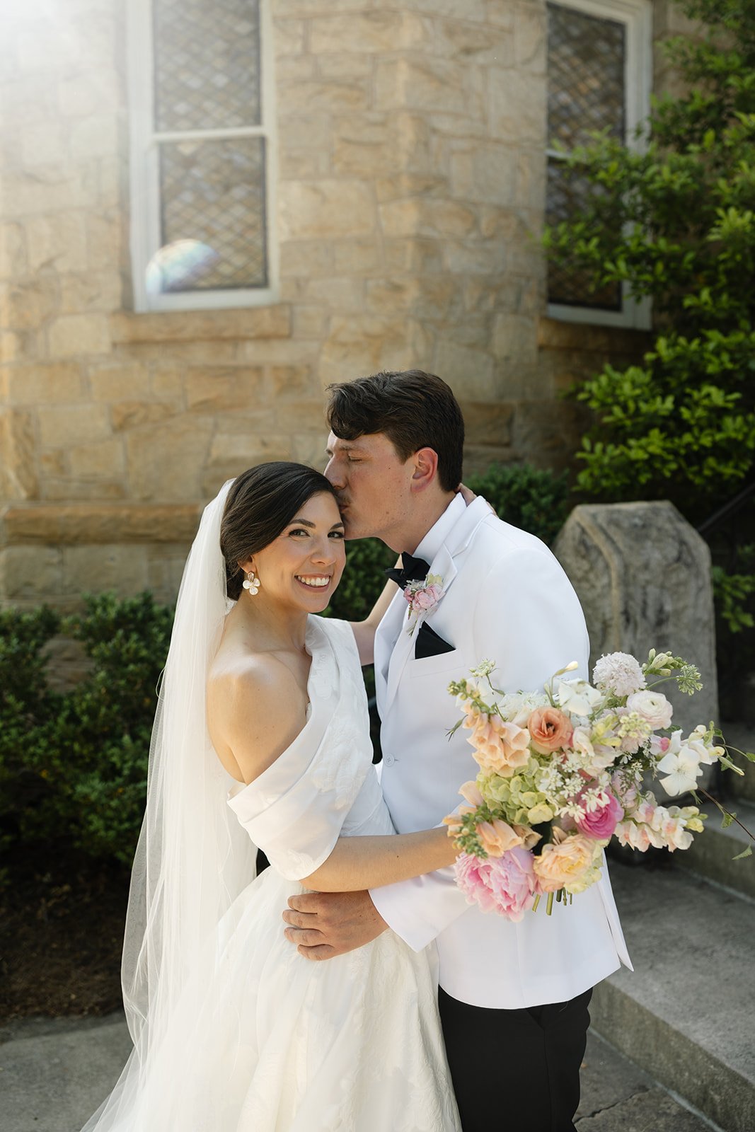 A newlywed couple in wedding attire embracing outdoors, with the groom kissing the bride on her forehead. The bride holds a bouquet of pink and white flowers, and the bride is smiling.