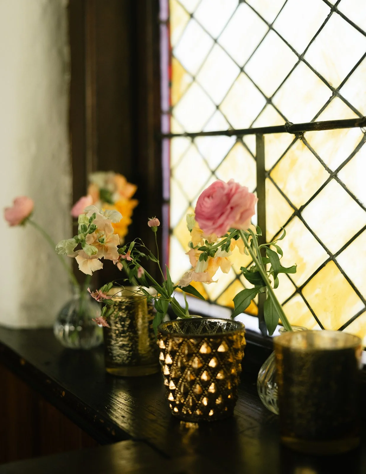 Vases with pink, white, and yellow flowers on a dark wooden surface near a stained glass window.