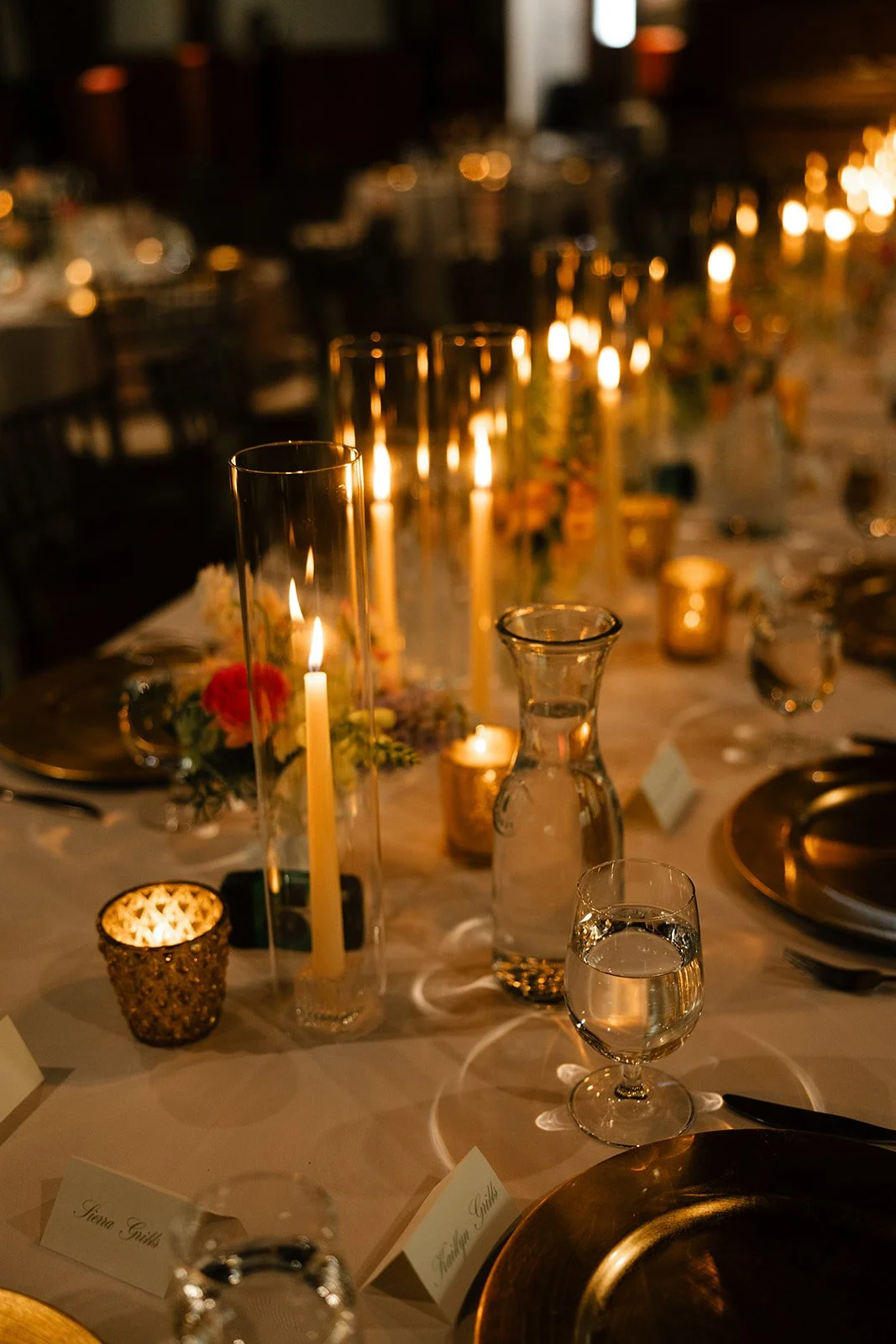 Lavishly decorated dinner table with tall candles, flowers, and glassware in warm candlelight at an elegant event.