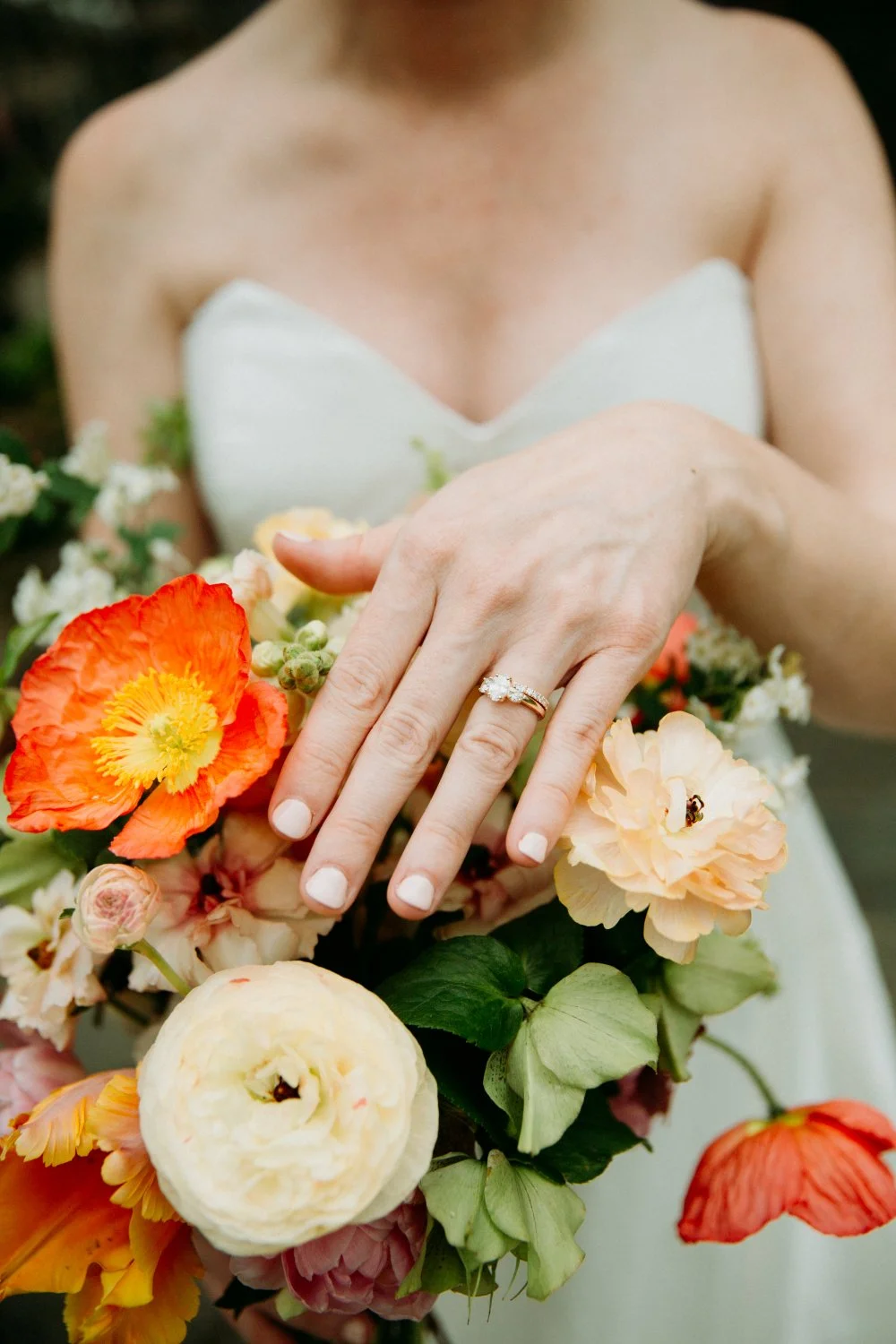 Close-up of a woman wearing a white strapless dress, showing her hand with a diamond engagement ring, holding a bouquet of orange, pink, white, and yellow flowers.