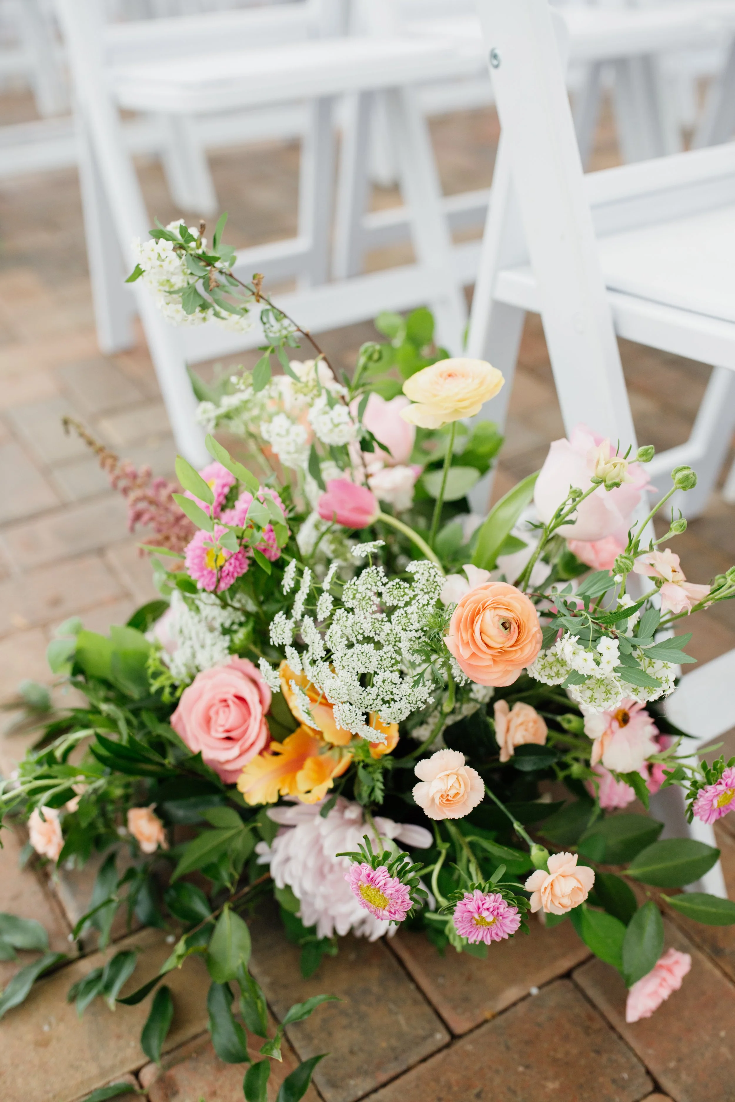 A colorful bouquet of various flowers placed on a brick floor with white chairs in the background.