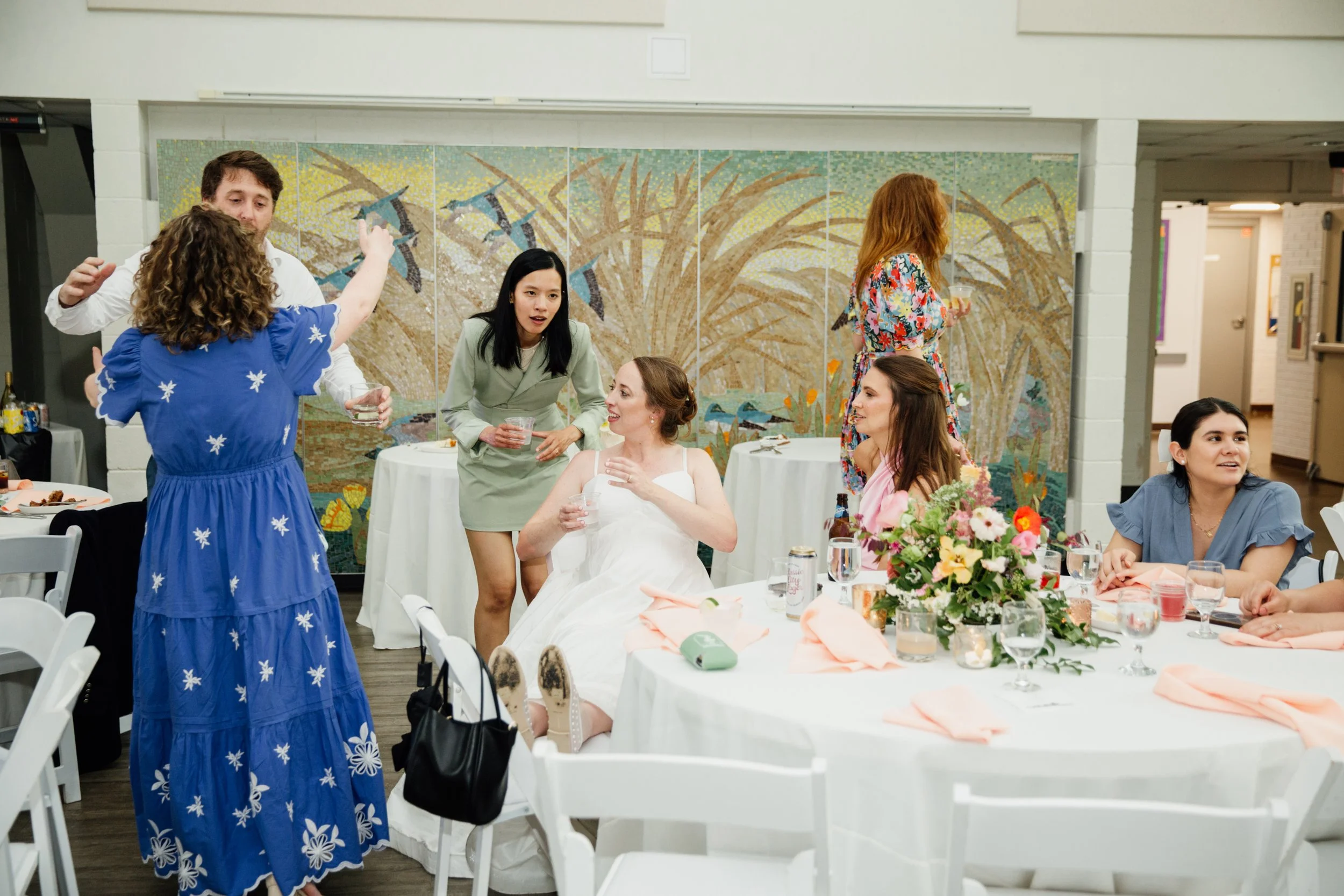 People at a wedding reception, some standing and some sitting around a table with a bride in a white dress, floral centerpiece, and pink napkins.