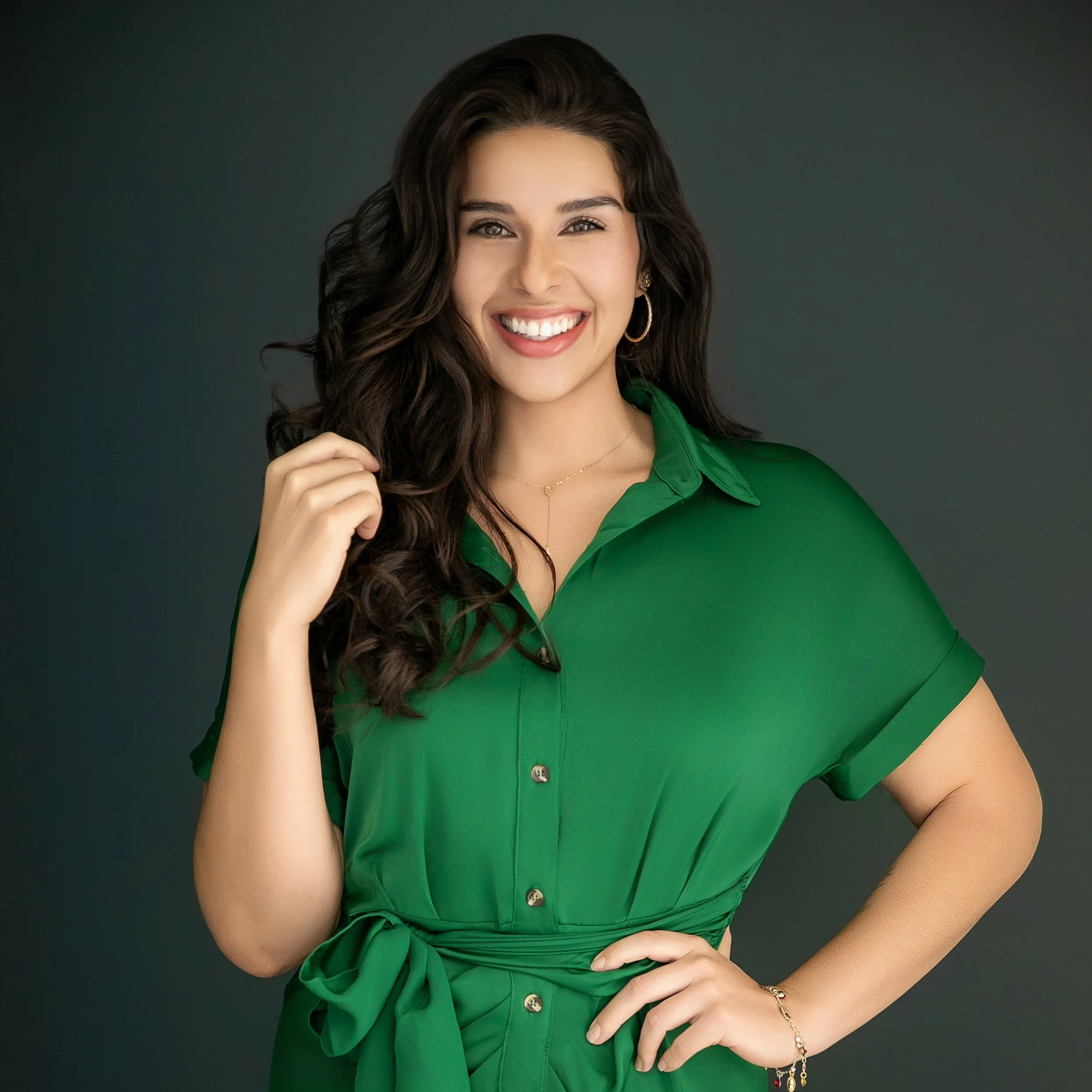 A woman with long, wavy dark hair, wearing a green dress with a waist tie, smiling and posing with one hand on her hip against a dark background.