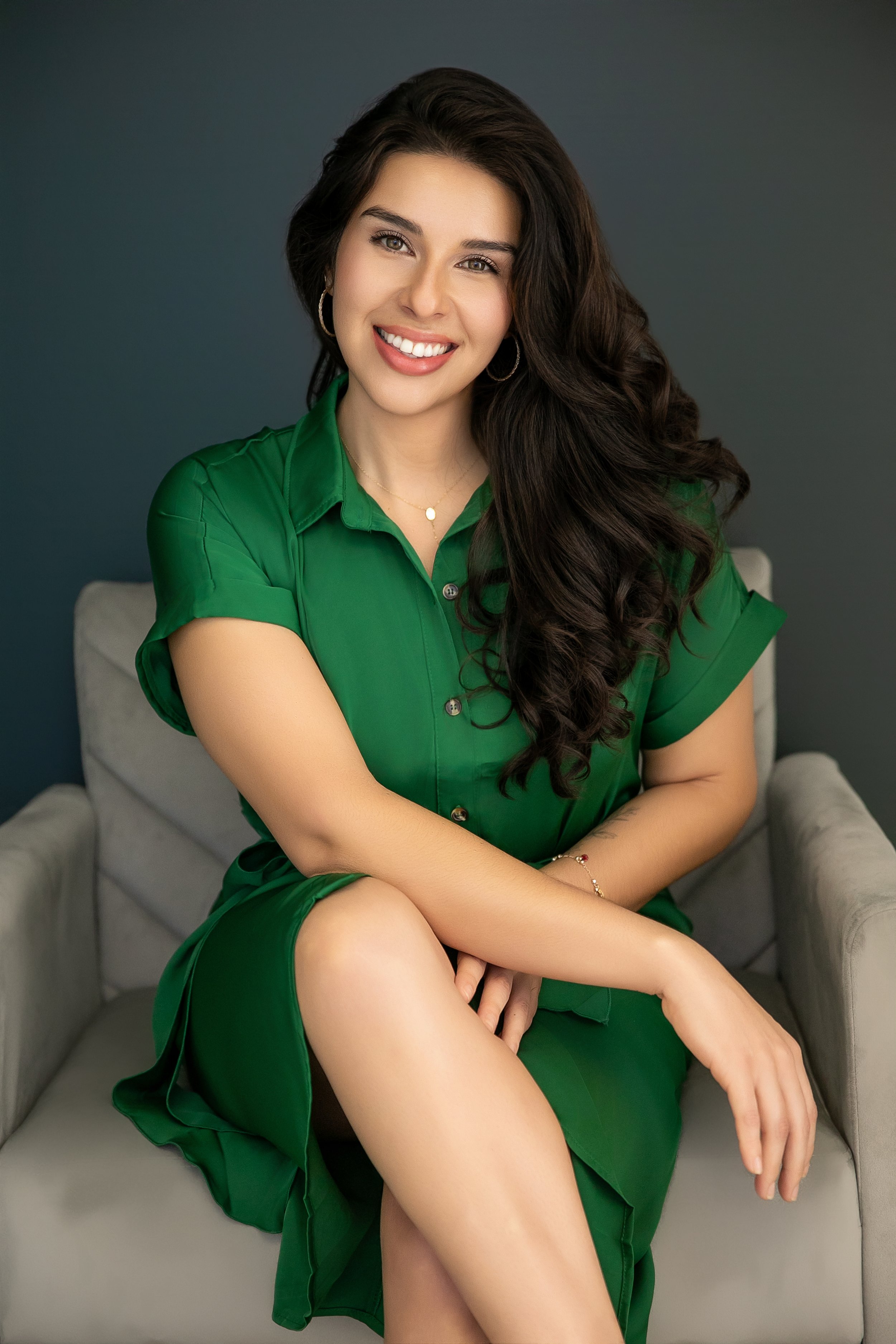 A woman with long dark hair, wearing a green dress, sitting on a beige couch, smiling at the camera against a dark grey background.