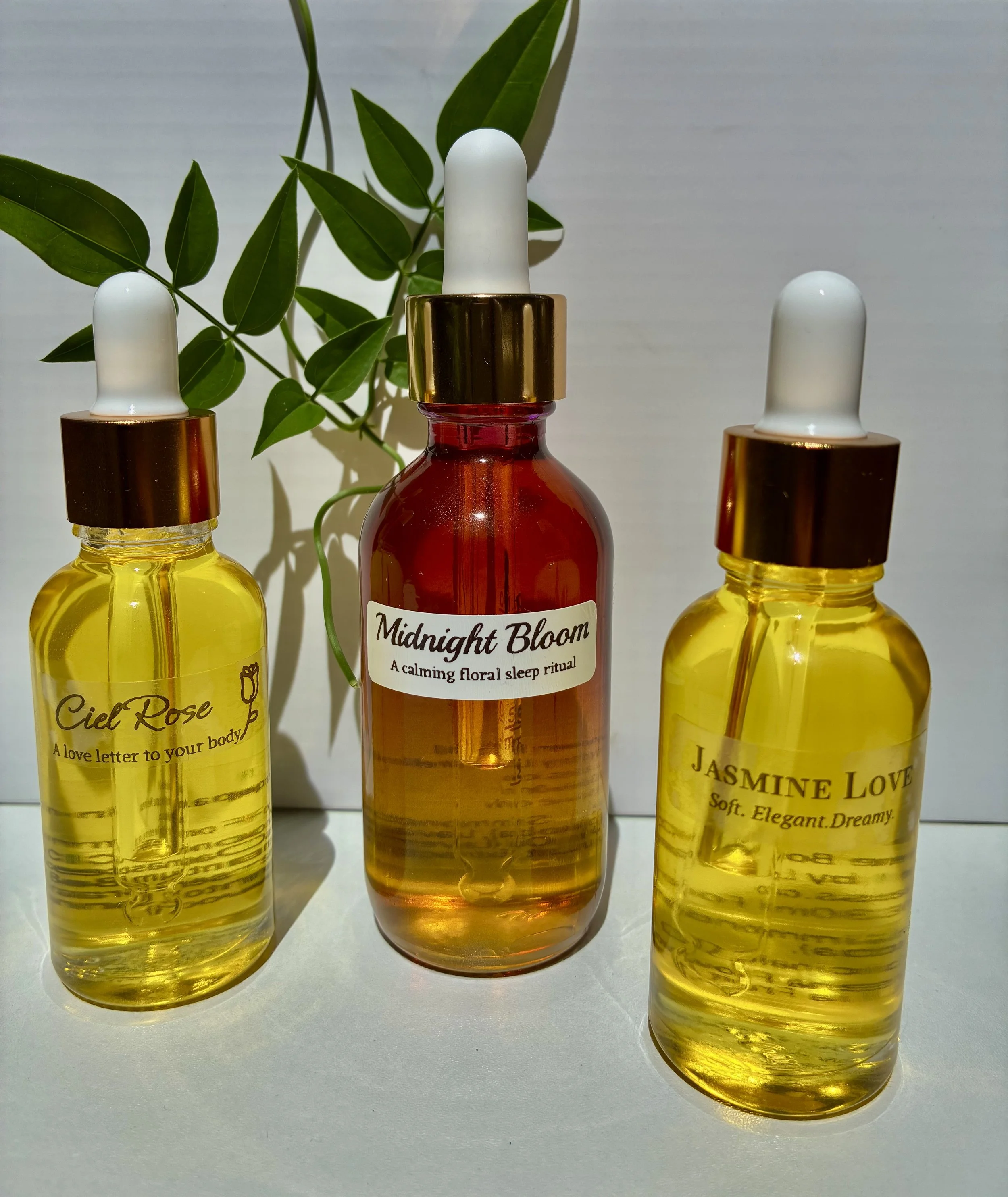 Three glass bottles with droppers, two yellow and one red, labeled 'Ciel Rose', 'Midnight Bloom', and 'Jasmine Love', with a leafy plant in the background.