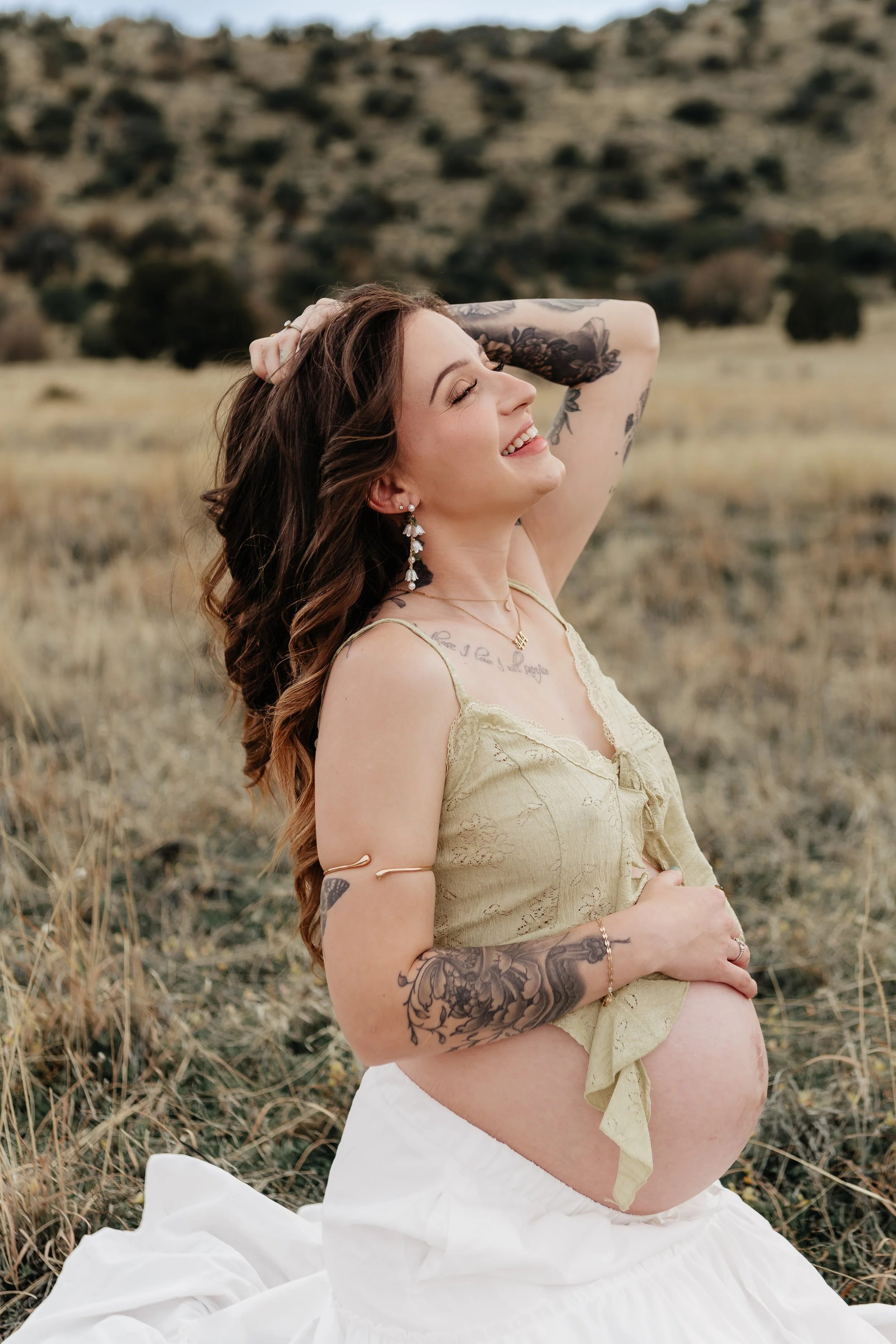 A pregnant woman with tattoos on her arms and shoulder, smiling with eyes closed and one hand on her head, kneeling outdoors on dry grass near a hill.