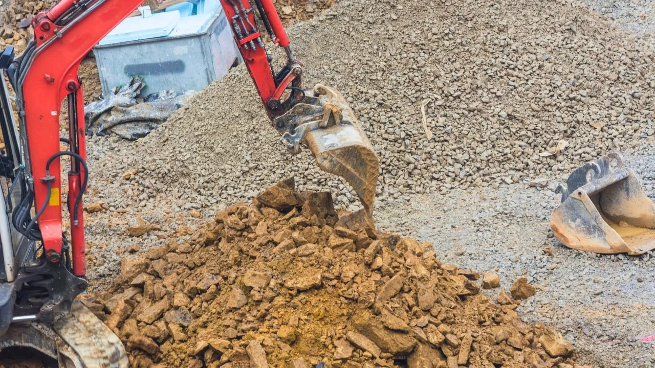 Excavator digging in dirt with piles of rocks and soil nearby.