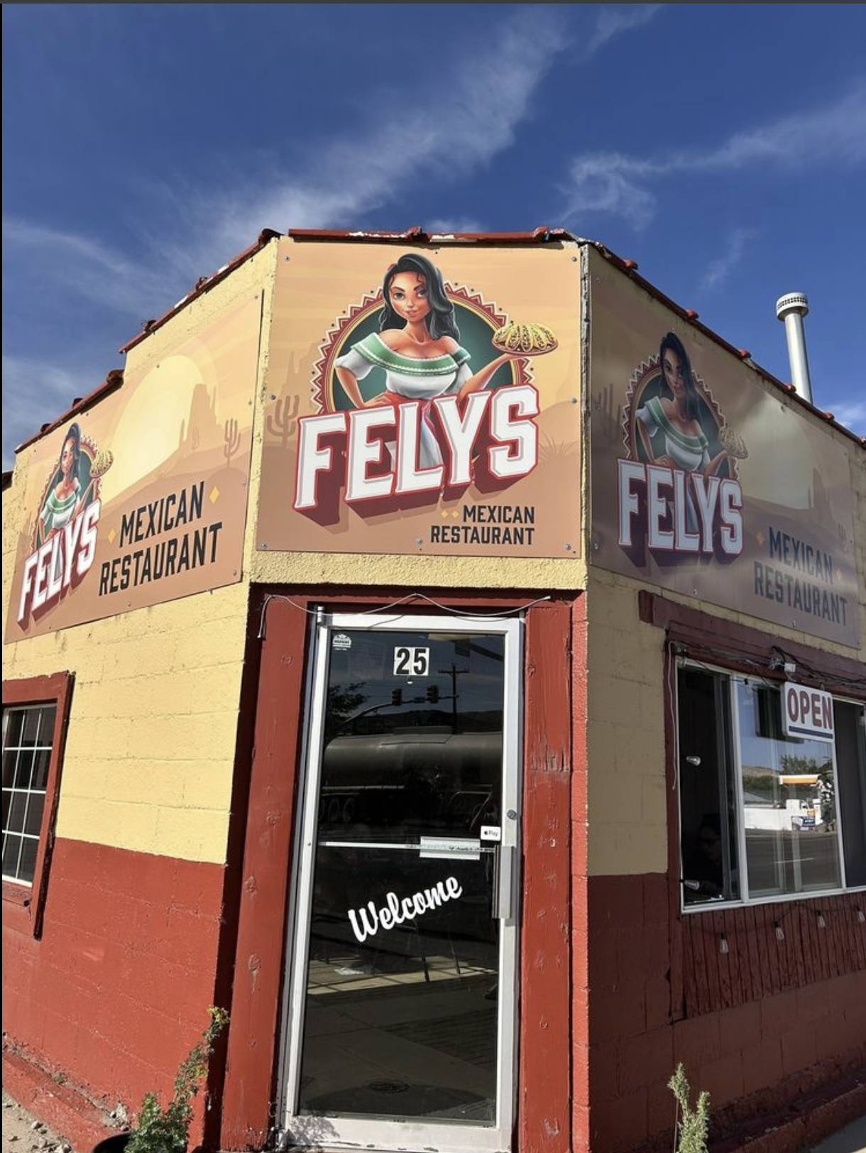 Exterior of Felys Mexican Restaurant with a large sign showing a woman in traditional Mexican dress holding a plate of food. The building has yellow and red walls, with an 'Open' sign in the window and a door with a 'Welcome' sign.