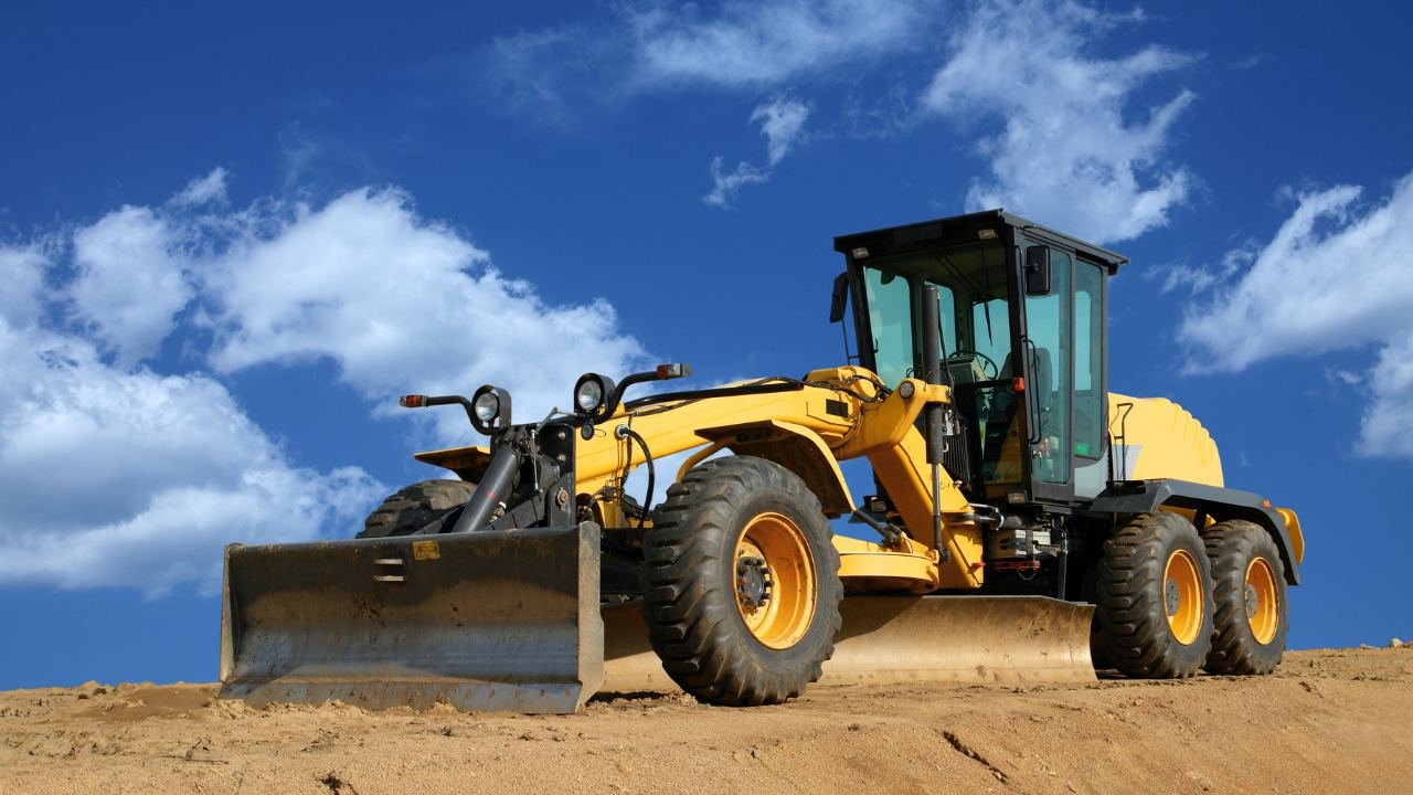 Yellow grader on sandy terrain with blue sky background.